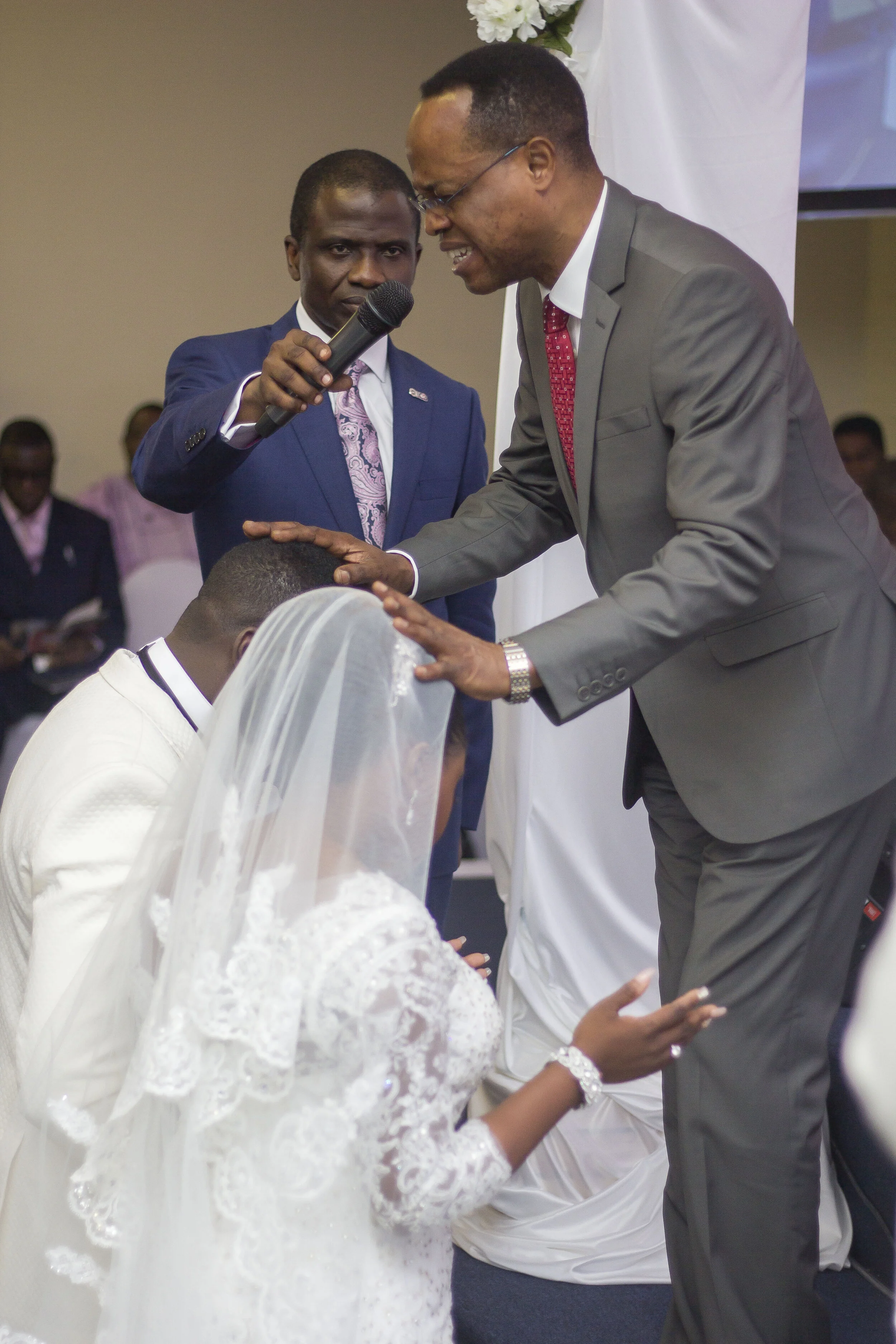 A bride and groom kneeling with their heads bowed during a wedding ceremony, surrounded by attendants including a man in a gray suit who is kneeling and touching the groom's head, and another man in a blue suit holding a microphone, with a white flor