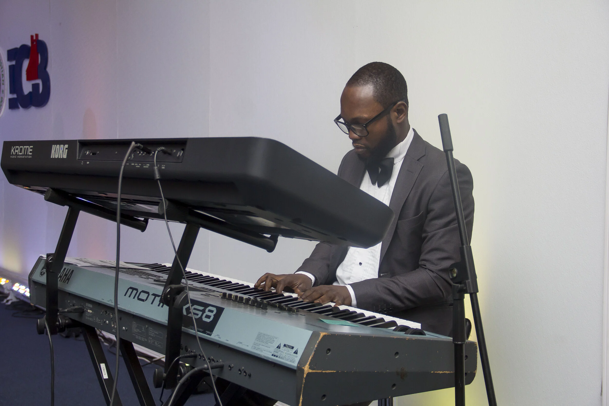 A man dressed in a tuxedo with a bow tie plays a keyboard. He wears glasses and has short hair and a beard. The keyboard is on a stand in front of him, and the wall behind him is plain and light-colored.