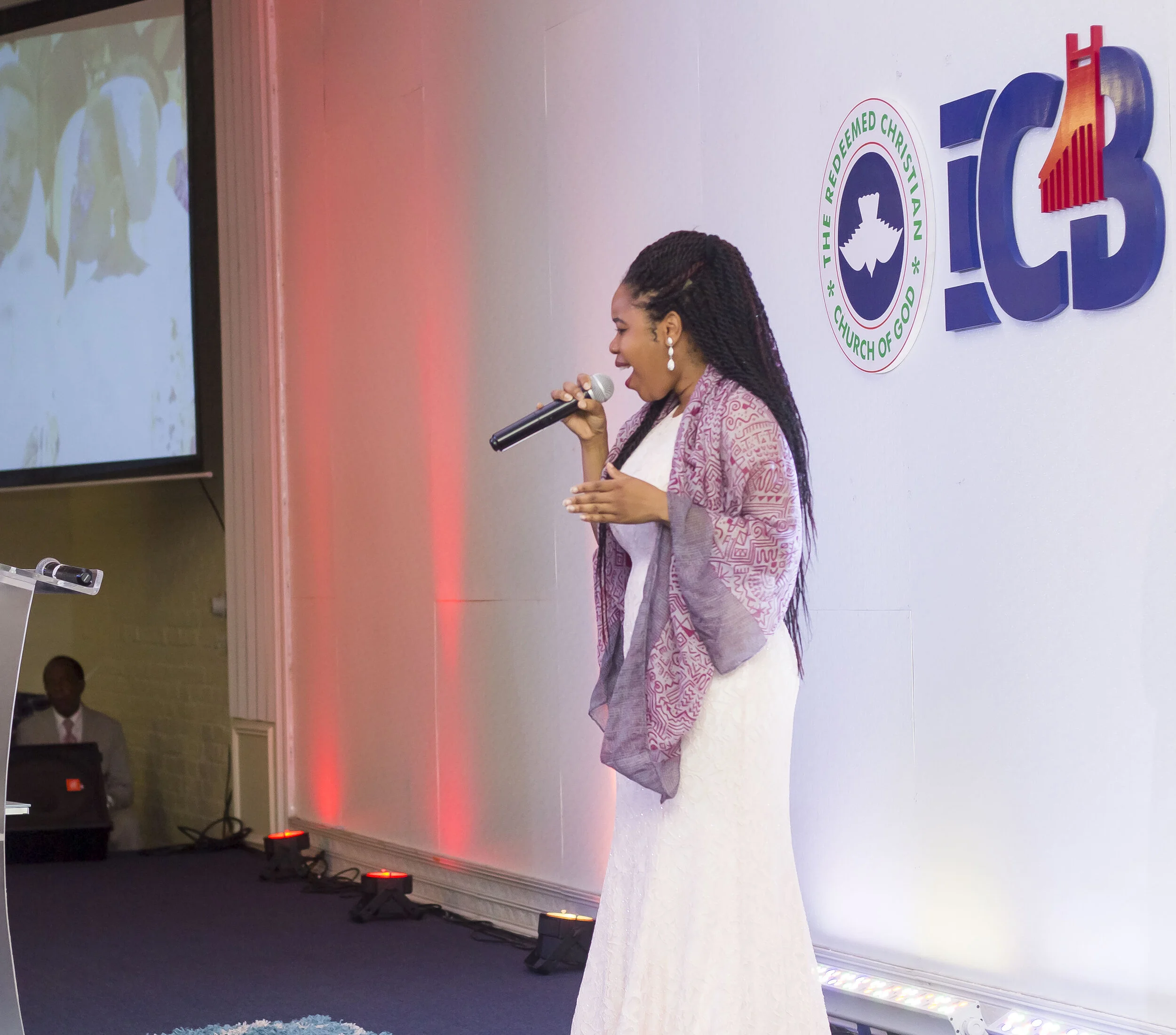 A woman with braided hair speaking into a microphone during an event at The Redeemed Christian Church of God, with a large sign displaying the church's logo and name on the wall behind her.
