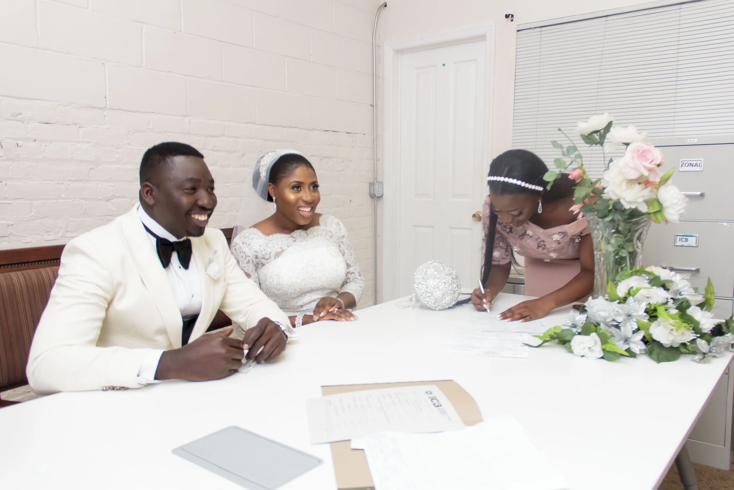 A bride and groom sitting at a table during a wedding ceremony, with a woman signing a document, and decorative flowers on the table.
