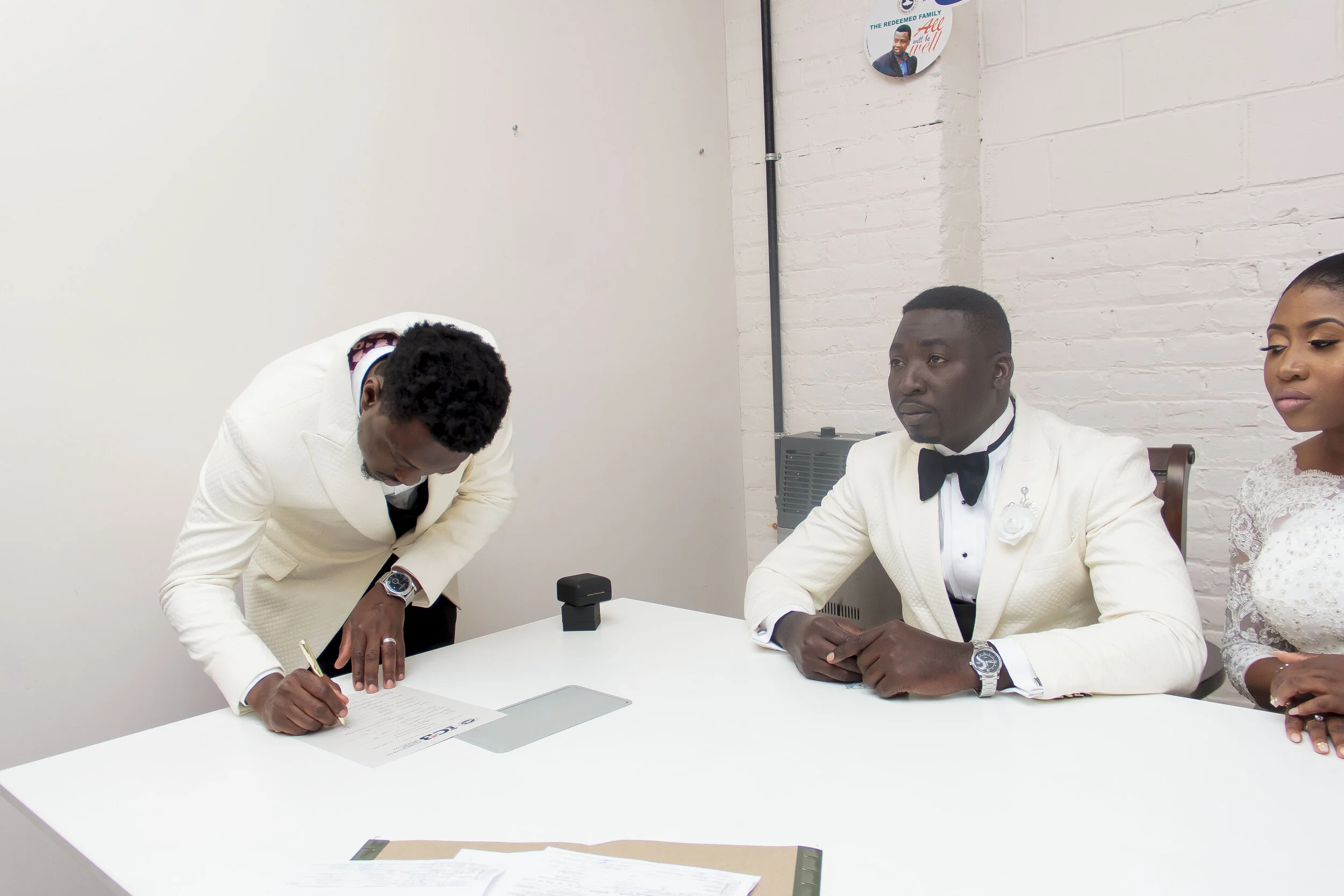 A man in a white tuxedo signs a document on a table, with two other people sitting nearby; the man is wearing a black bow tie, and the woman next to him is in a white lace dress.