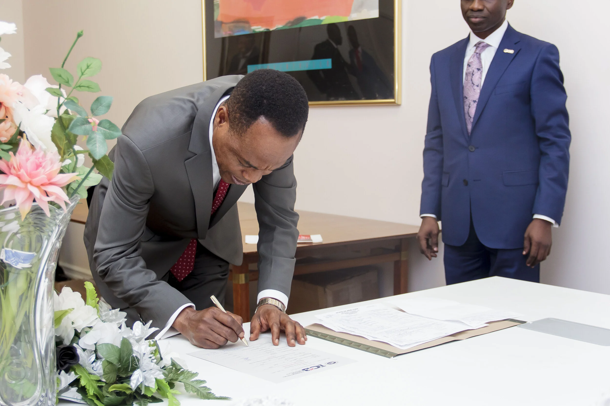 A man in a gray suit signing documents at a table with a woman in a blue suit standing nearby in an office. The table has paperwork and a bouquet of pink and white flowers.