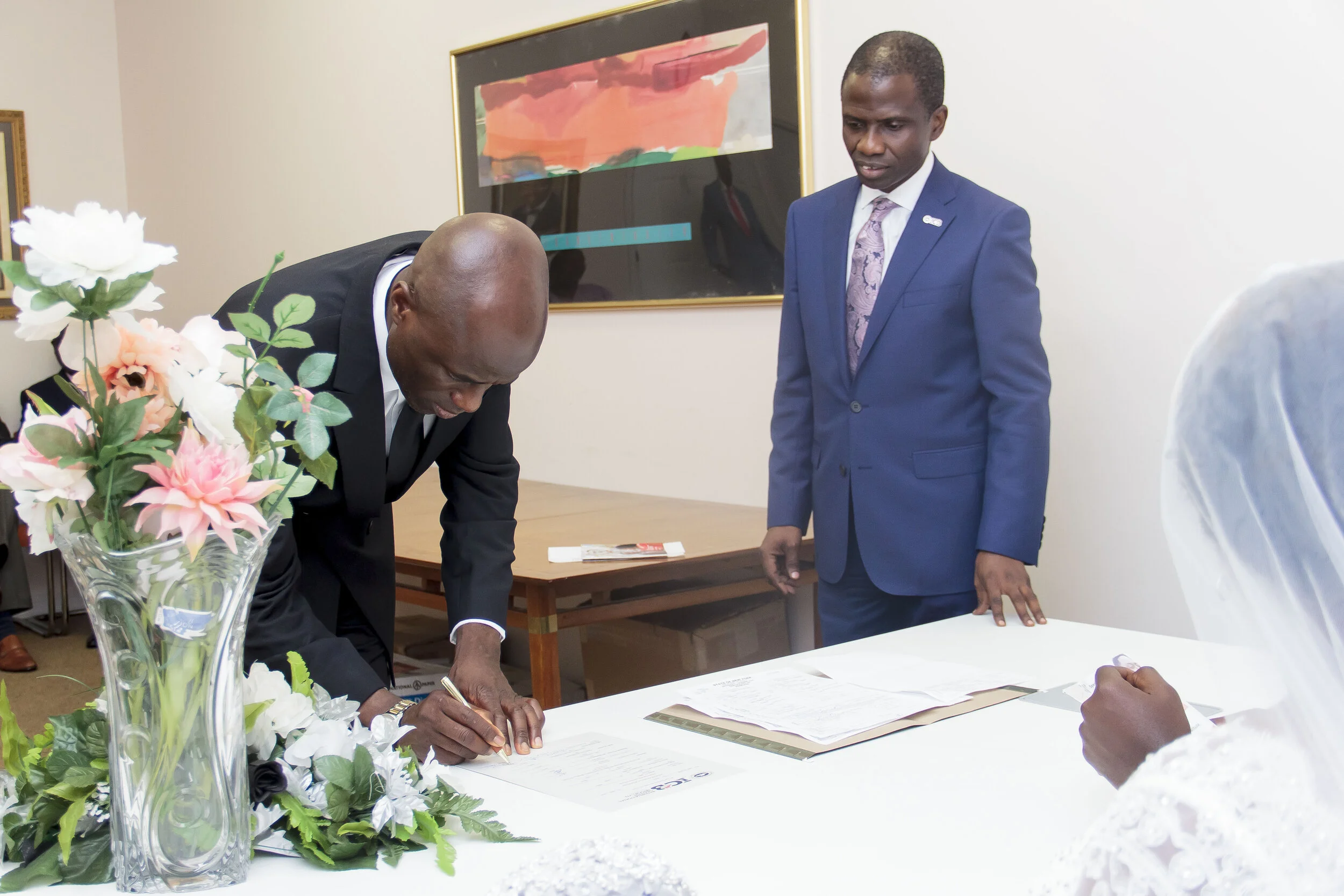 A man in a dark suit and a man in a blue suit looking at documents on a table, with flowers in a vase in the foreground and a colorful abstract painting on the wall behind them.