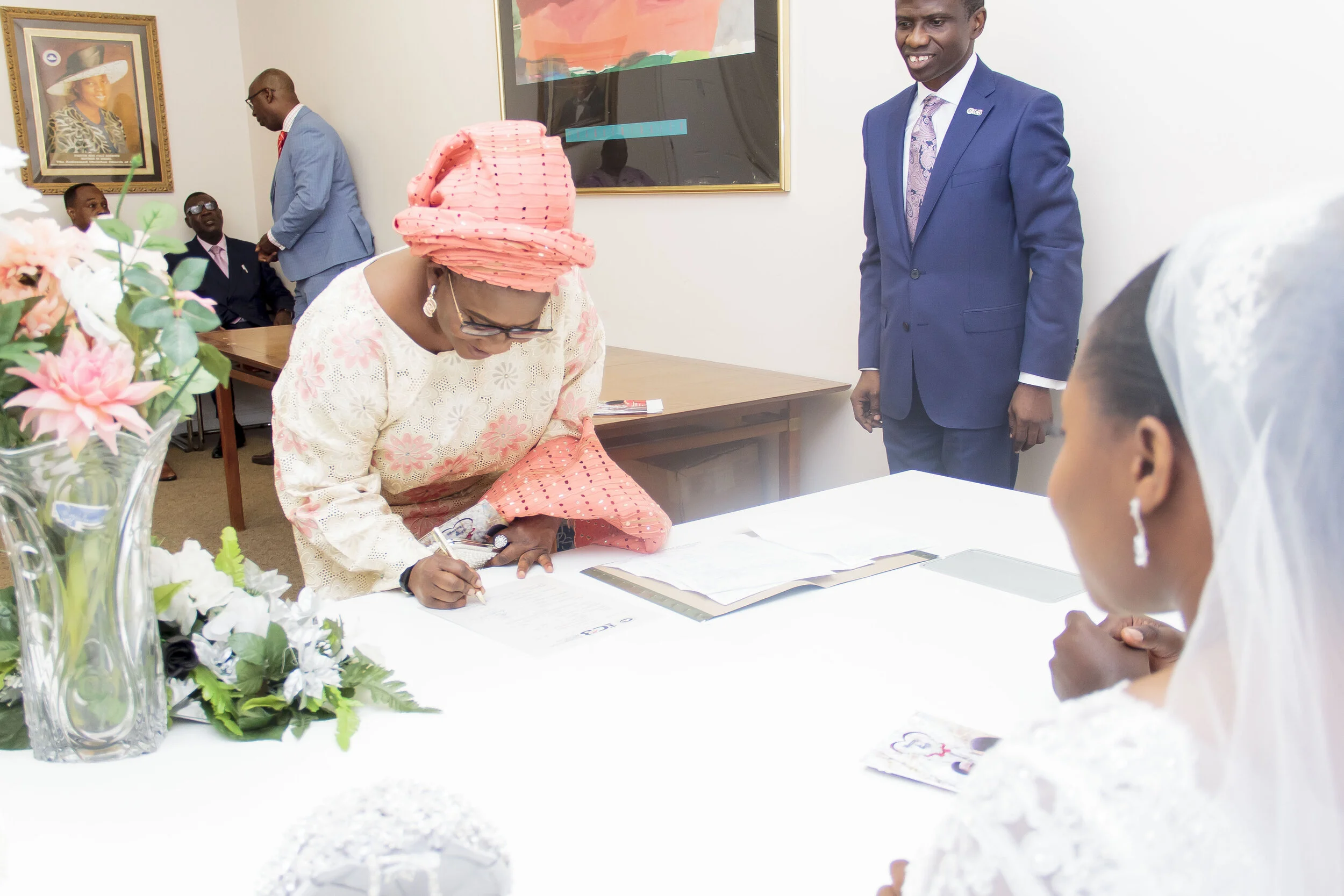 A woman in traditional African attire, including a pink headwrap and glasses, signs a document at a formal event. Several other people are in the room, some seated and some standing, with a large floral arrangement on the table.