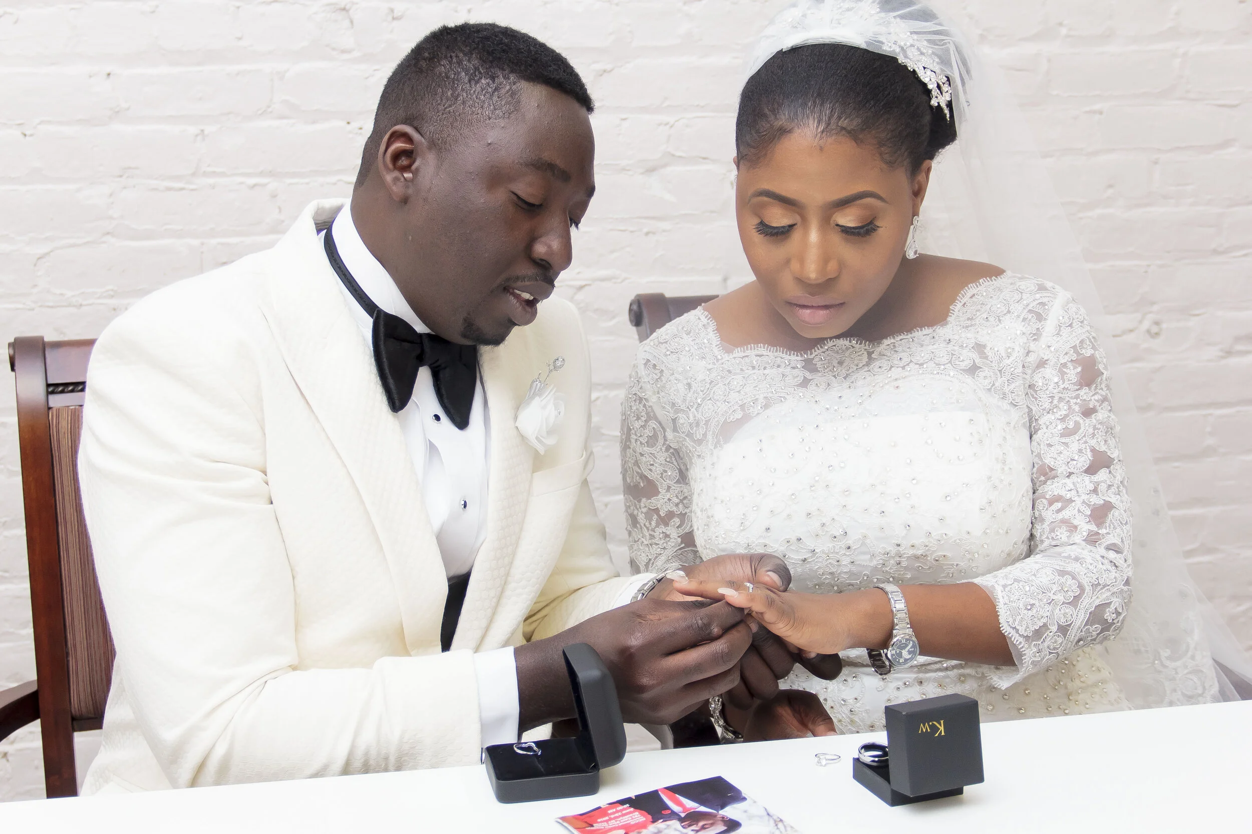 A newlywed couple at their wedding ceremony sitting close with the groom holding the bride's hand and looking at her. They are dressed in formal wedding attire, with the groom in a white tuxedo and the bride in a white lace wedding dress with a veil.