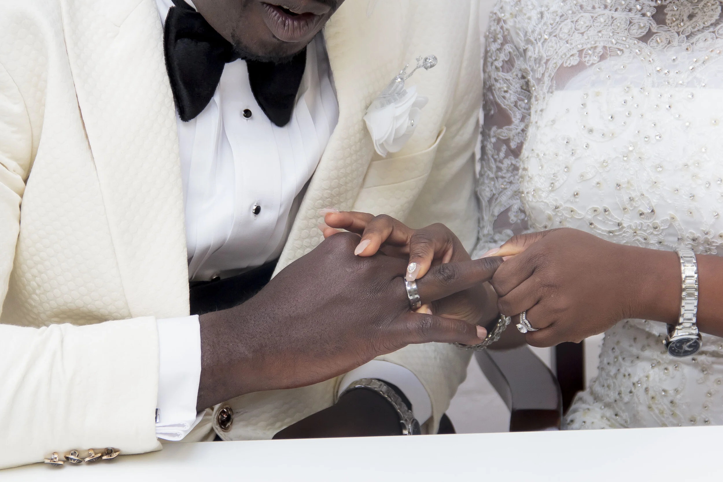 A couple dressed in wedding attire holding hands, with the woman wearing a white lace wedding dress and the man wearing a cream-colored tuxedo with a black bow tie. They are clasping hands with wedding rings visible.