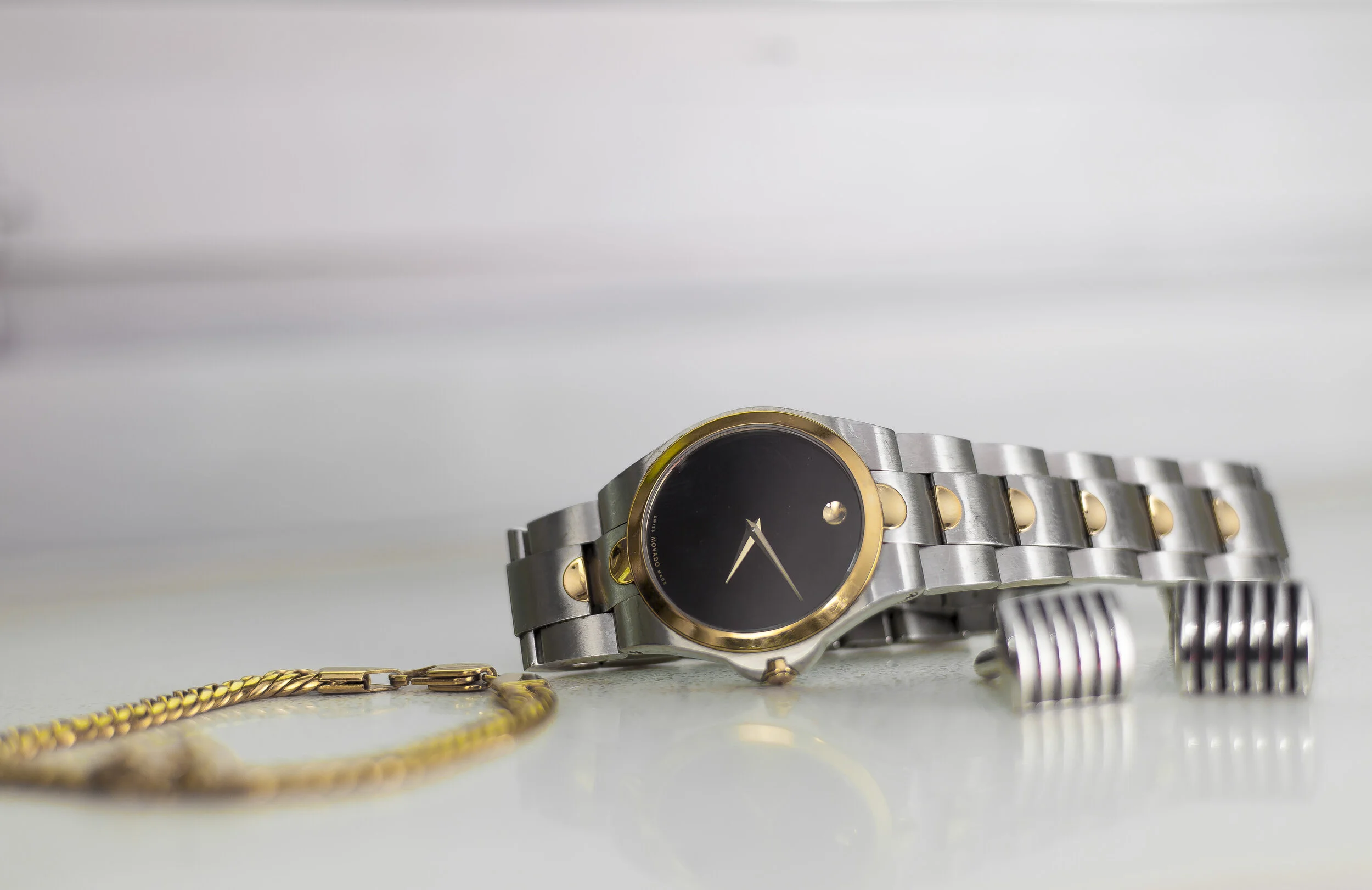 A wristwatch with a black face, gold bezel, and a silver and gold metal band, resting on a white surface.