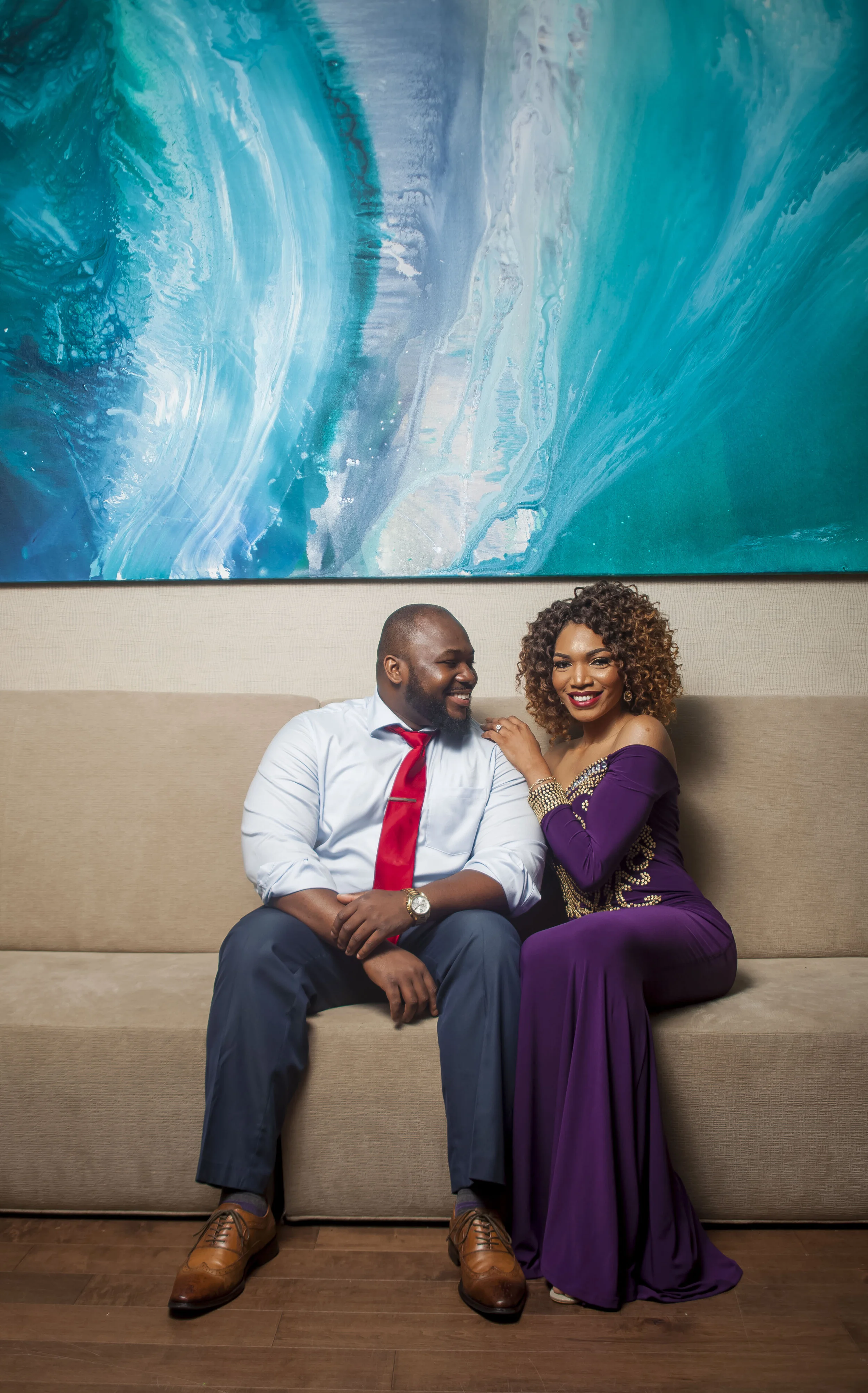 A man and a woman sitting close on a beige couch, smiling and engaging with each other. The man wears a white shirt, red tie, dark pants, and brown shoes. The woman wears a purple dress with gold embellishments and has curly hair. Behind them is a la
