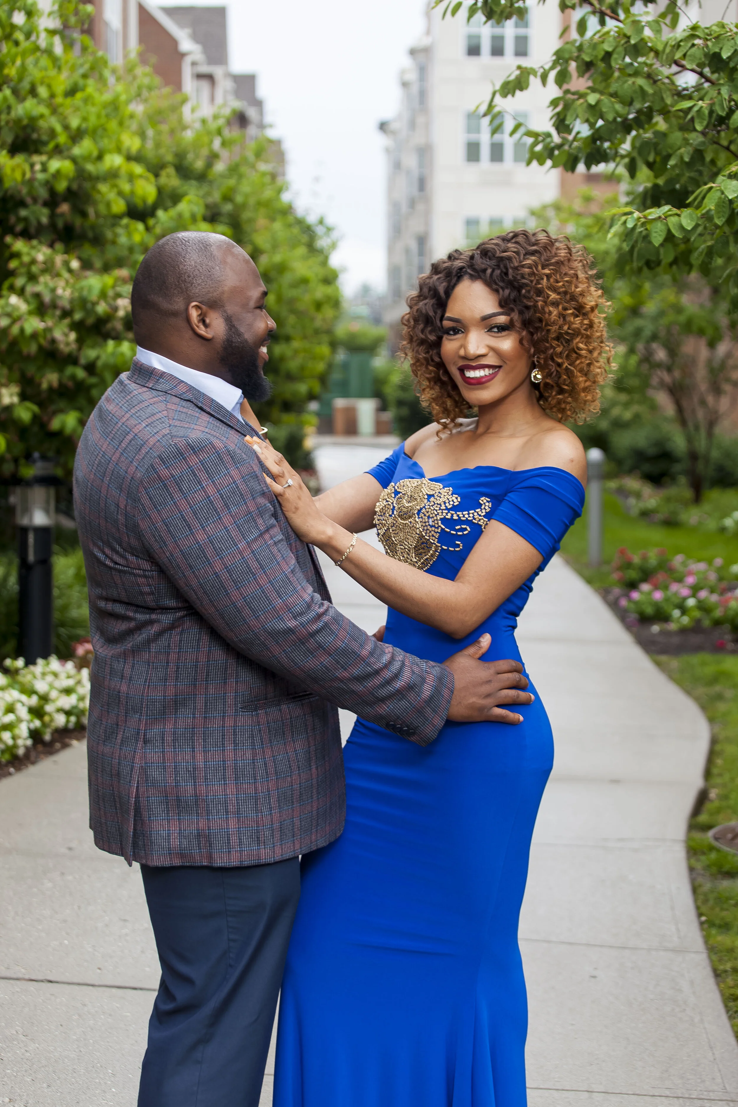 A smiling woman in a blue dress with gold embroidery holding hands with a man in a plaid blazer outdoors in a neighborhood with greenery and apartment buildings.