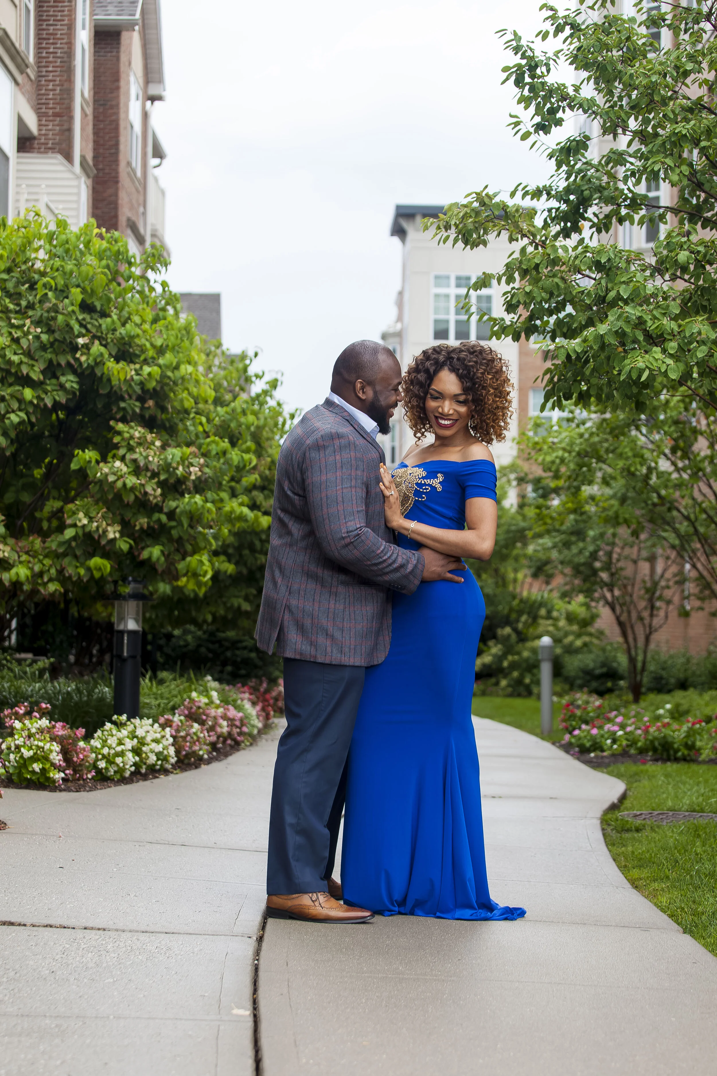 A couple dressed elegantly, smiling, and holding each other outdoors on a sidewalk lined with flowers and trees in a residential neighborhood.