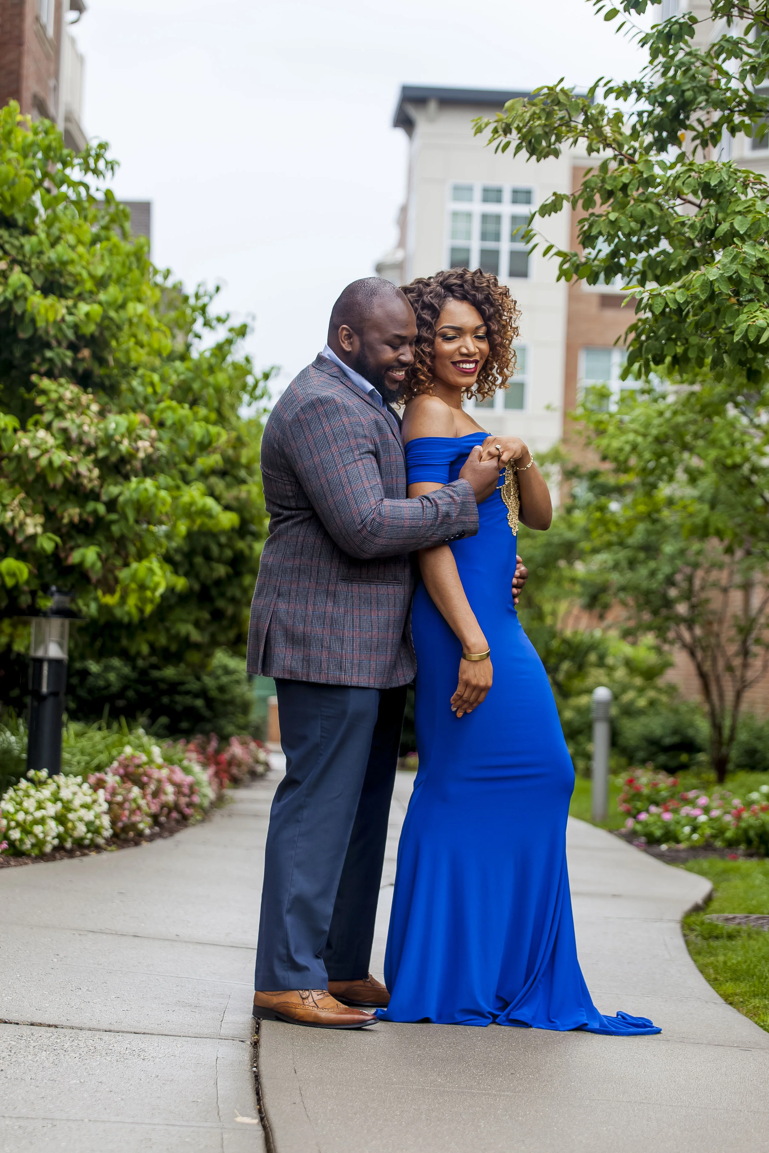 A couple dressed in formal attire standing on a sidewalk in a park-like setting with trees and flowers, smiling and embracing each other.