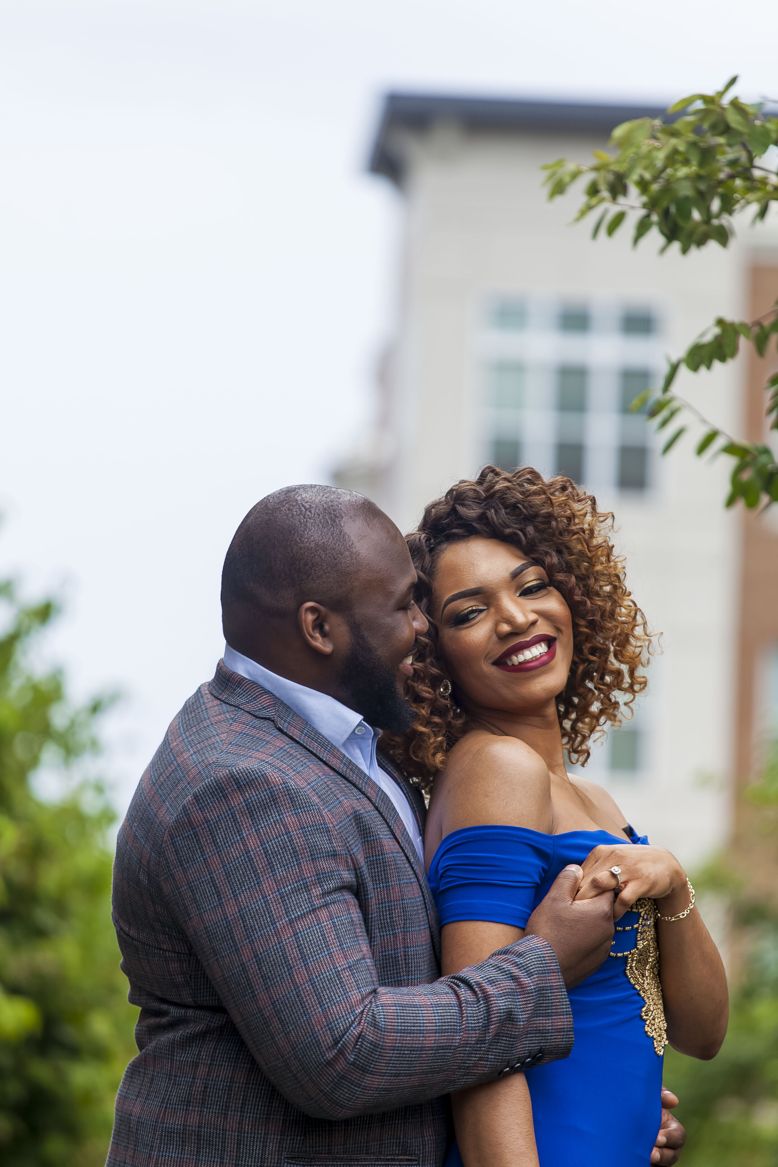 A happy couple hugging outdoors, with a woman in a blue dress and a man in a patterned blazer, with a residential building and greenery in the background.