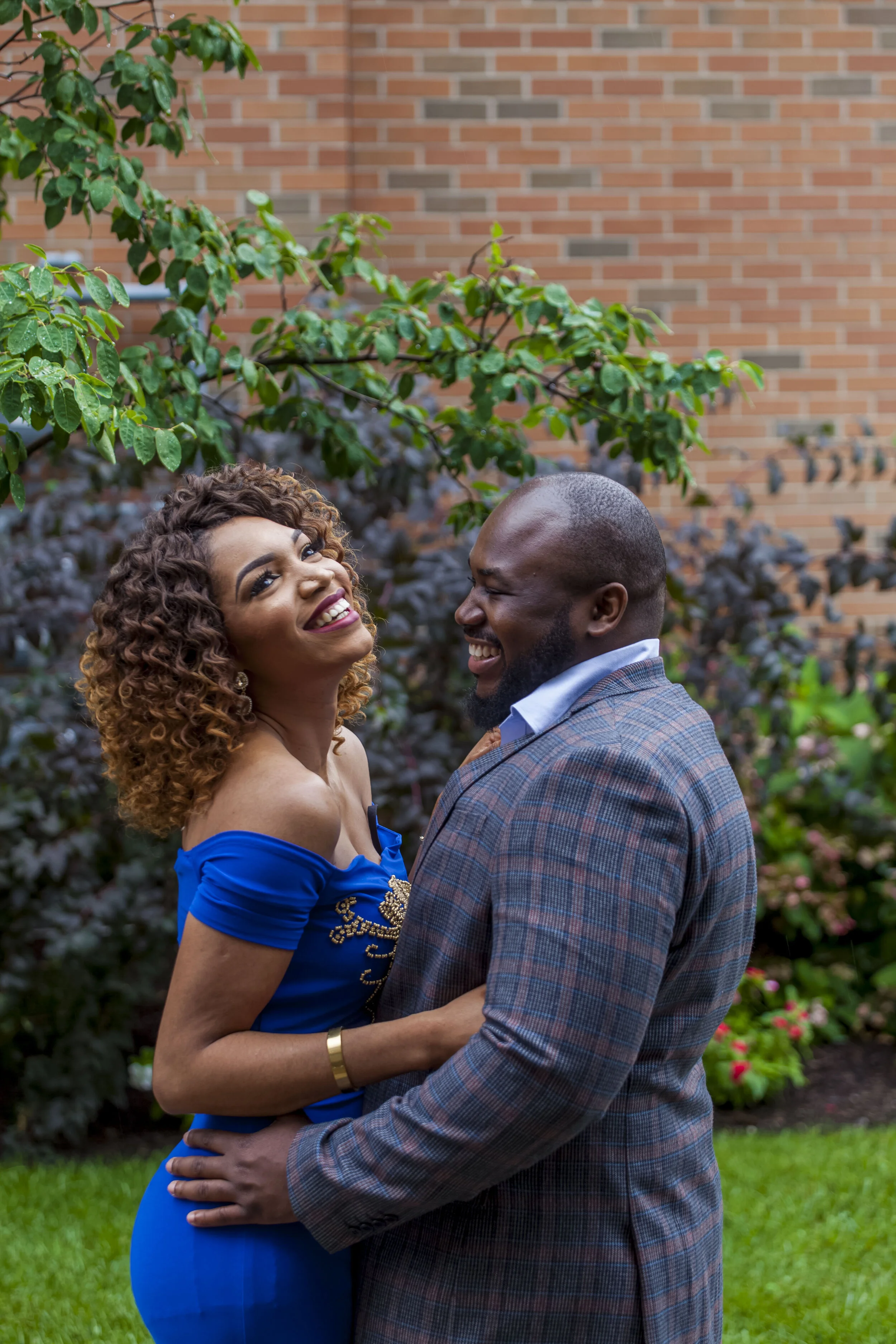 A smiling woman with curly hair wearing a royal blue off-shoulder dress with gold embellishments, and a gold bracelet, is being embraced by a man with a beard, dressed in a plaid suit jacket and a light blue shirt, in a garden with green foliage and 
