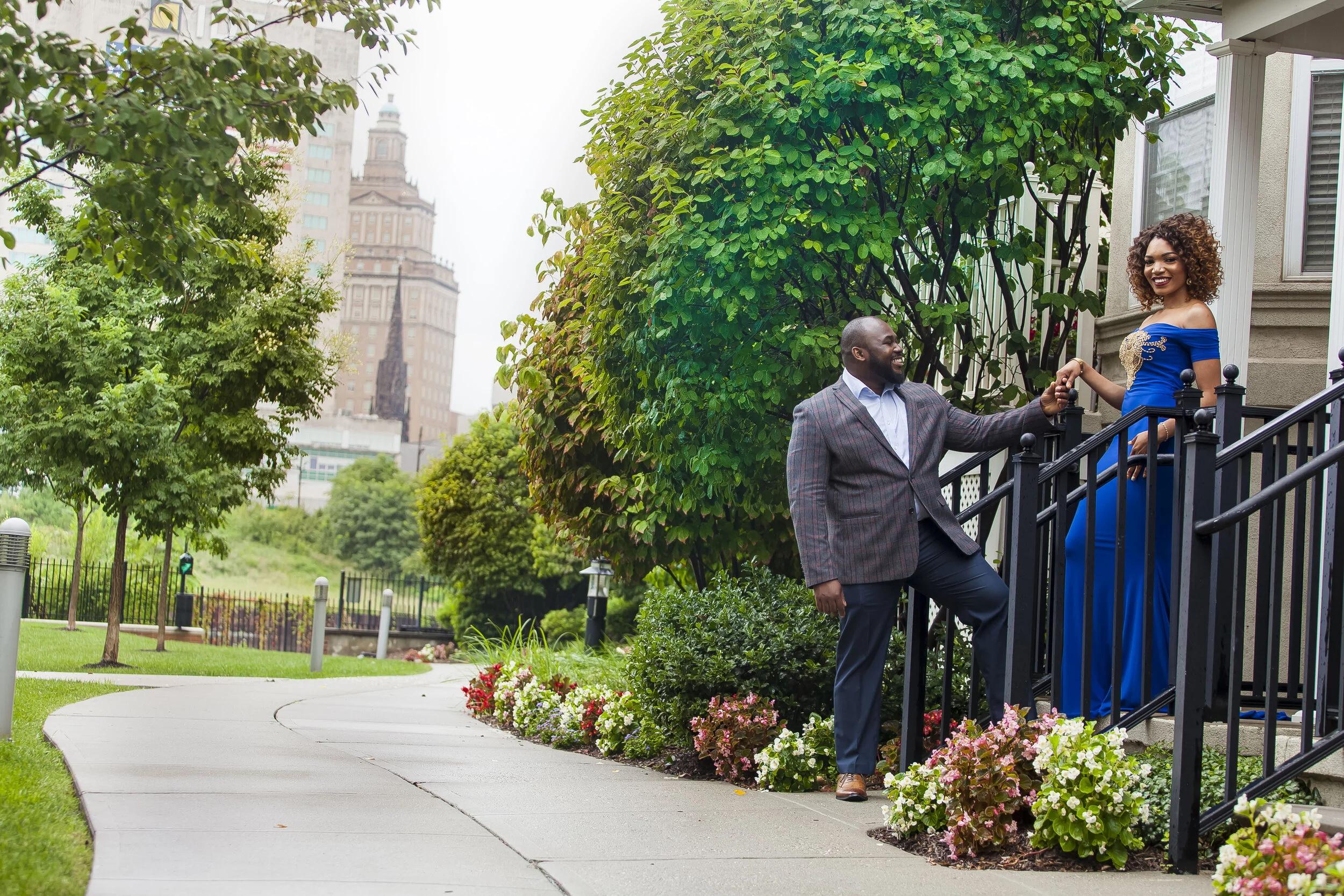 A man and a woman smiling and holding hands on the front steps of a house in an urban park setting with trees, flowers, and a city skyline in the background.