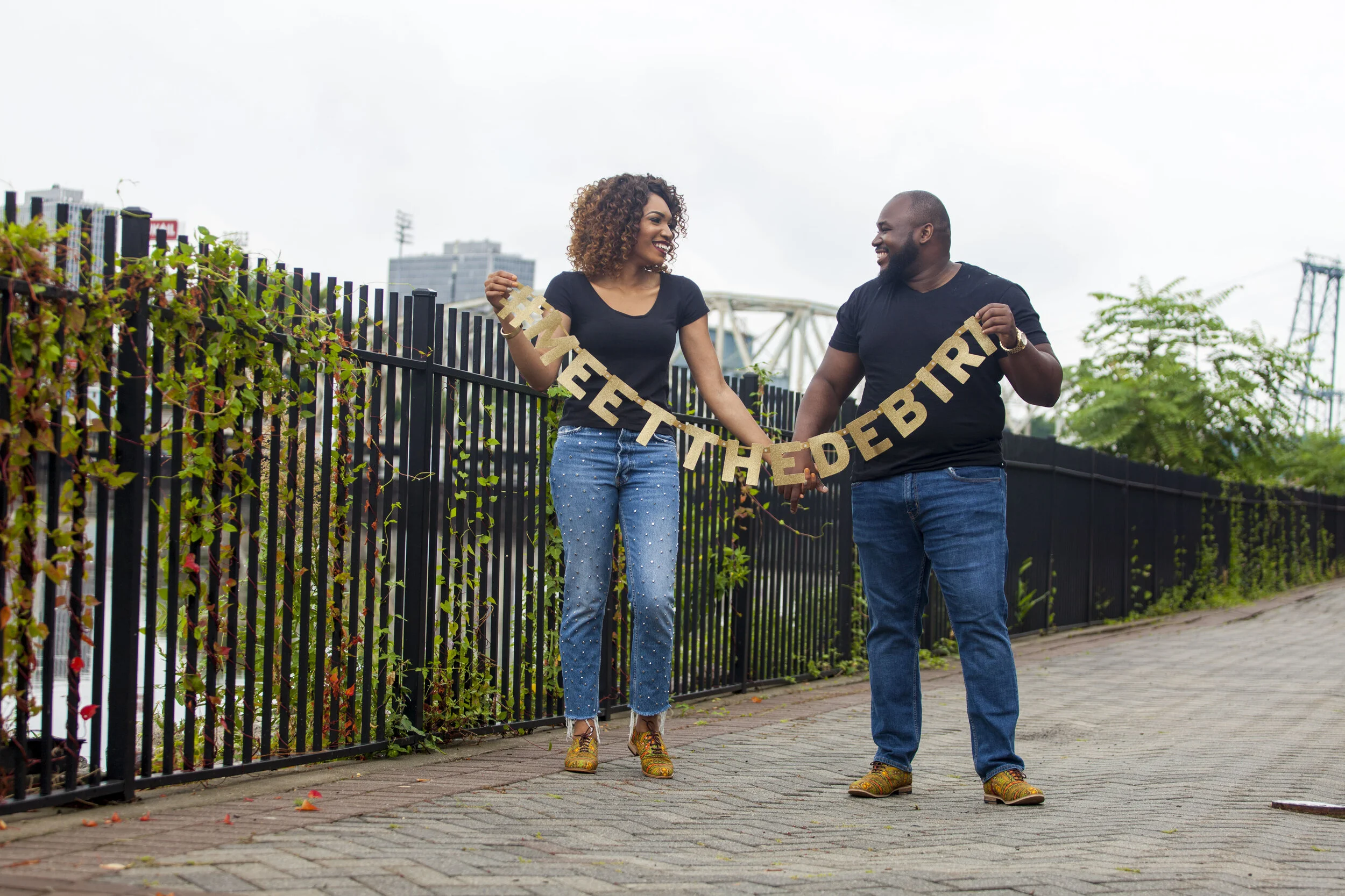 A man and woman celebrate a birthday outdoors, holding a gold 'Happy Birthday' banner and smiling at each other, with a city skyline in the background.