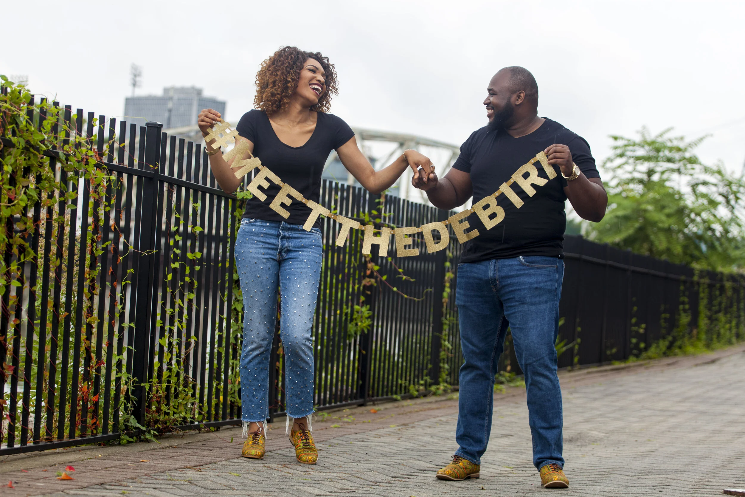 A woman and a man are outdoors, holding a gold glittery 'Happy Birthday' banner, smiling and joyfully dancing together. The woman has curly hair and is wearing a black t-shirt, blue jeans with pearl embellishments, and colorful shoes. The man has a b