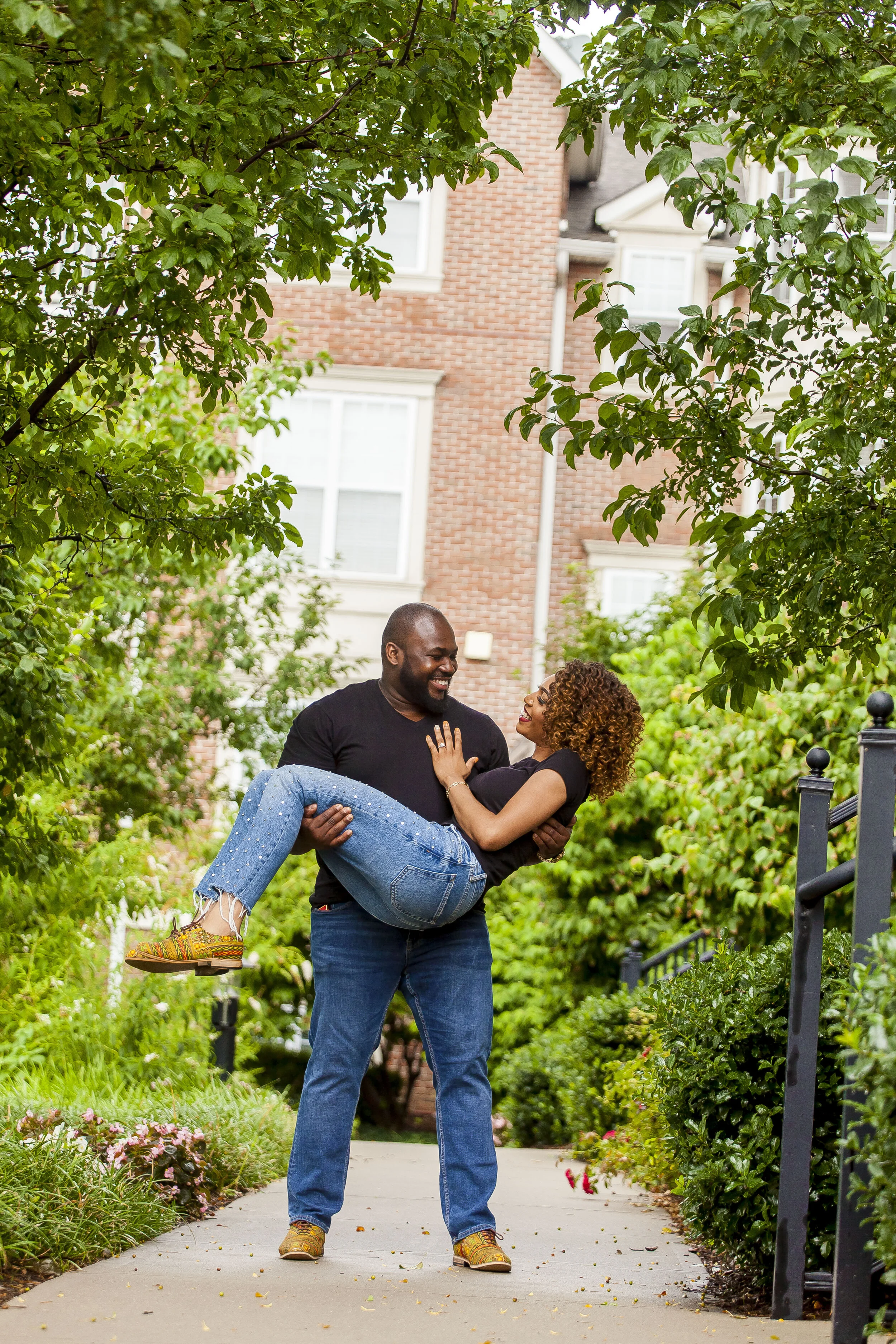 A man is carrying a woman in his arms while they smile at each other outdoors on a sidewalk with greenery and trees around. There is a brick building in the background.