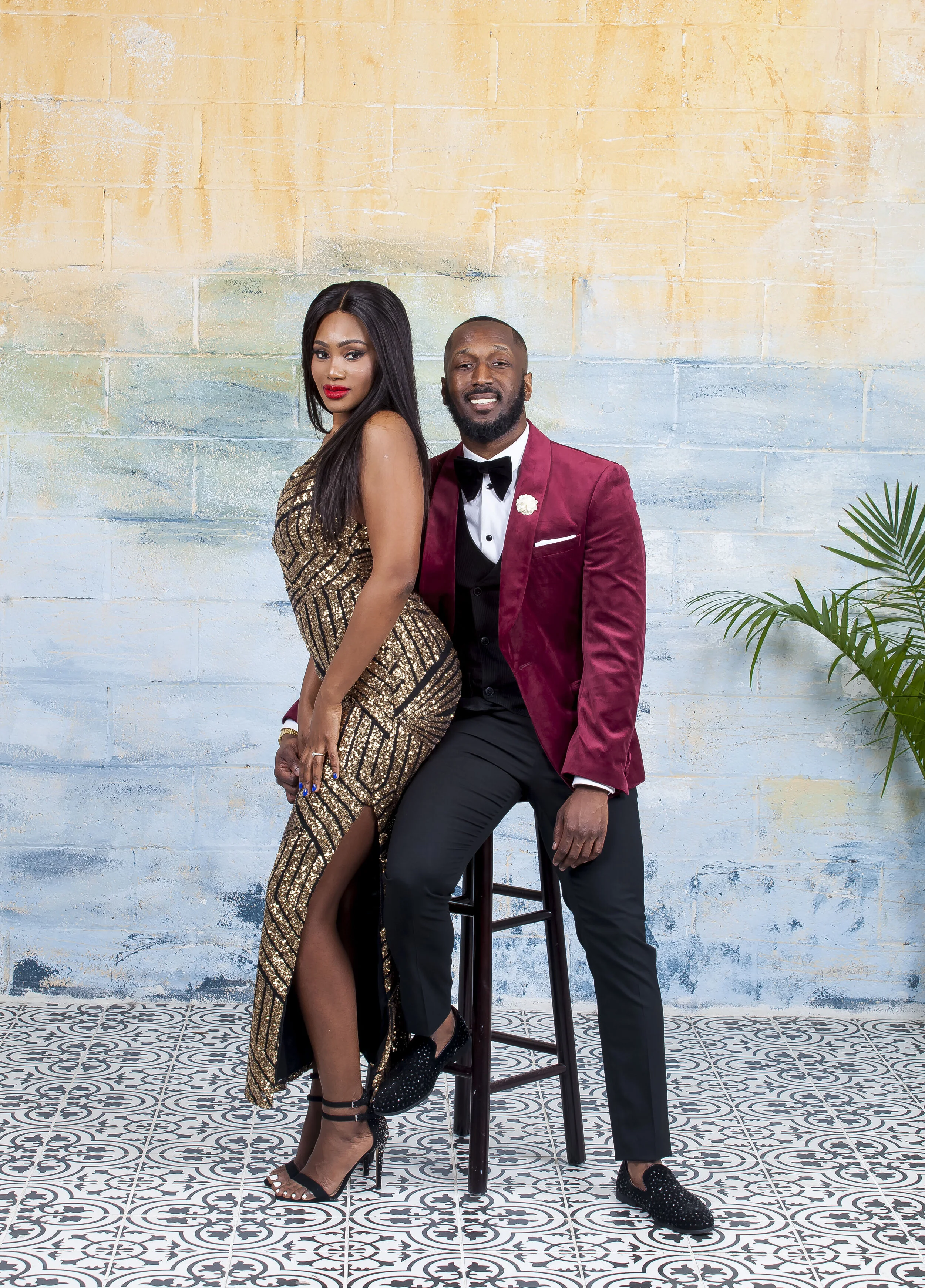 A man and woman dressed in formal attire standing in front of a textured, multicolored wall with a plant to the right. The woman is wearing a gold, sequined, slit dress and high heels. The man is wearing a burgundy velvet blazer, black trousers, and 