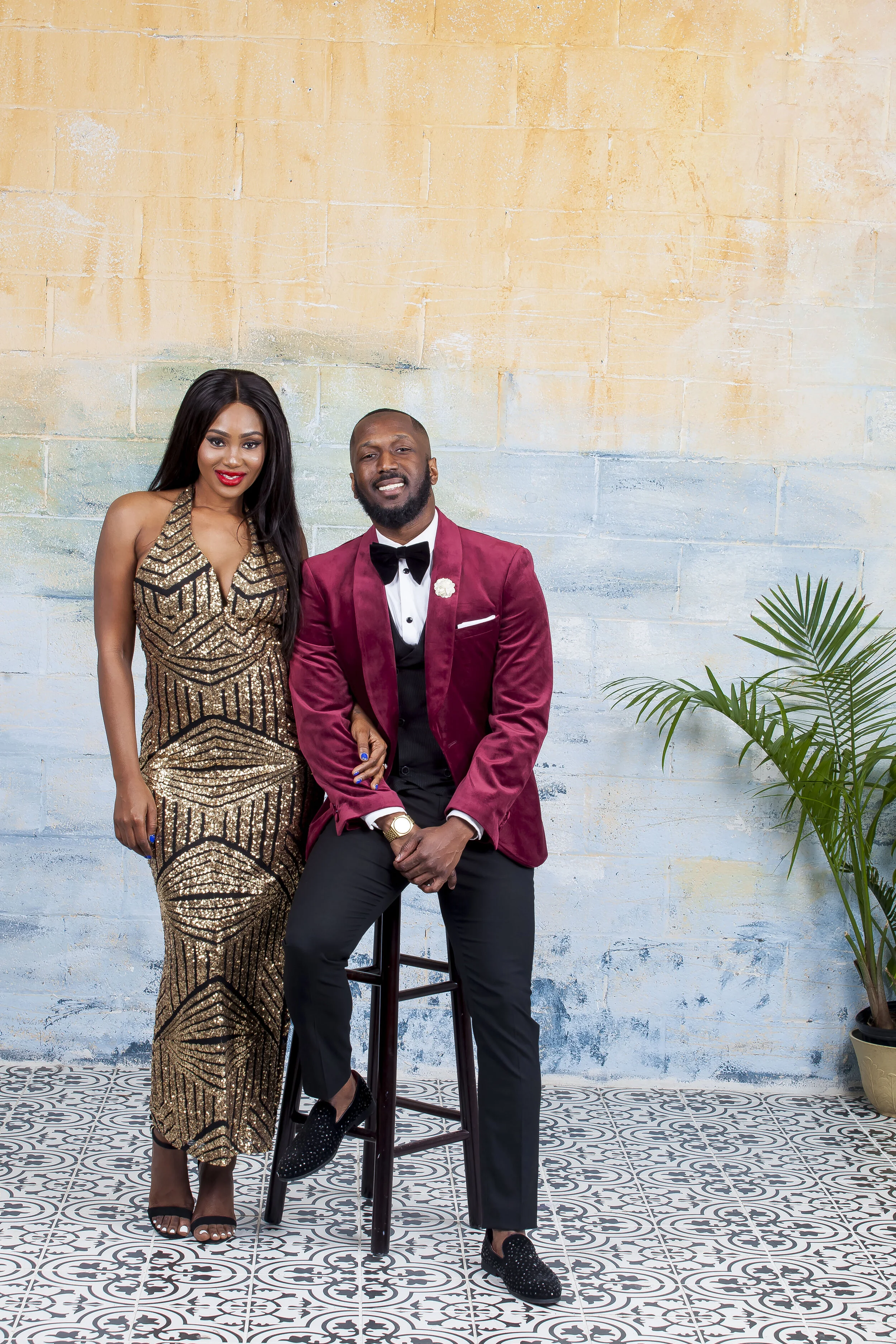 A stylish African-American couple dressed in formal attire posing against a painted brick wall with a potted plant on the right. The woman wears a gold and black sequined gown, and the man is in a red velvet blazer, black trousers, and a bow tie.