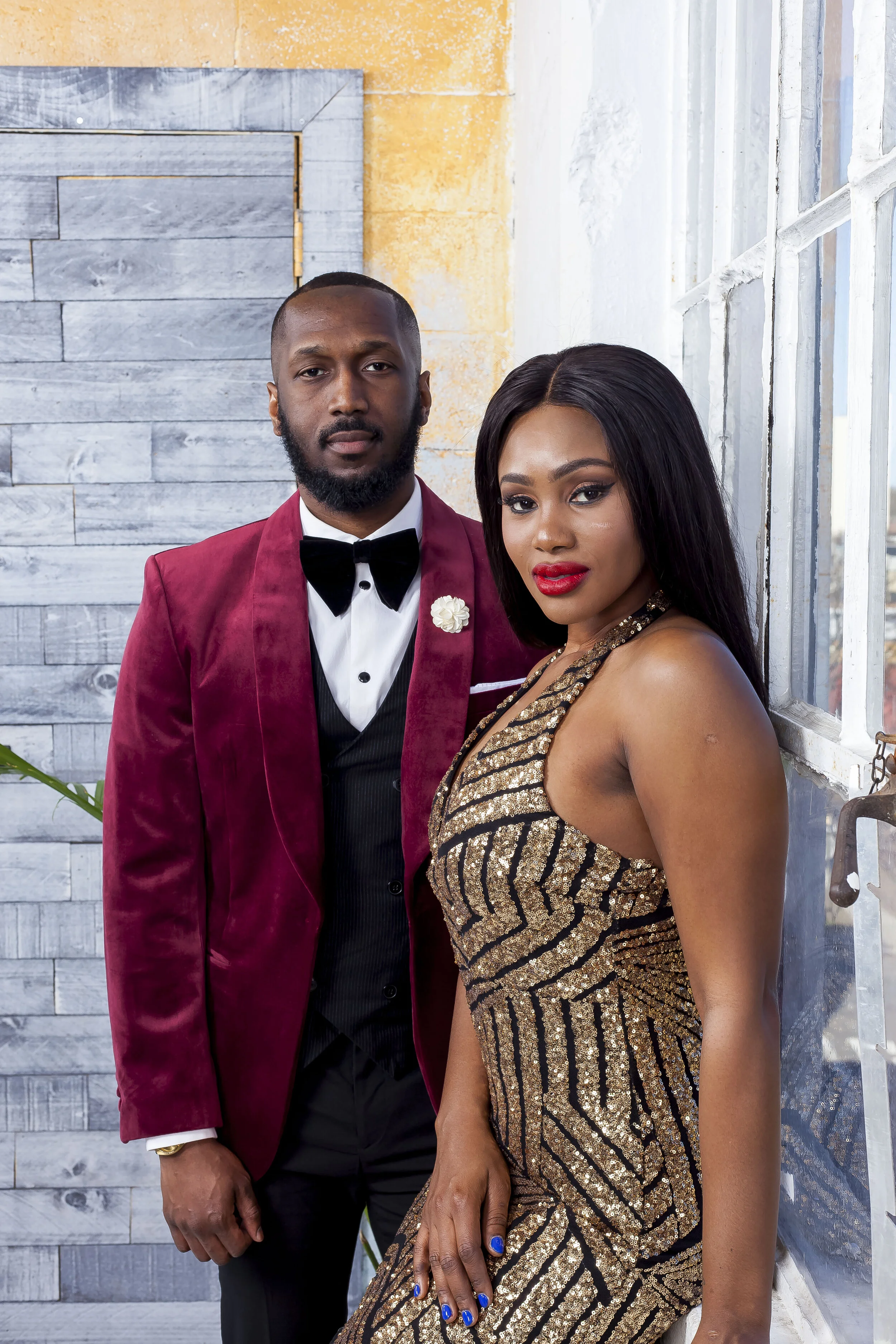 A man in a red tuxedo jacket with a black bow tie and a woman in a gold and black sequined dress posing together against a background with white-paneled windows and a yellow wall.