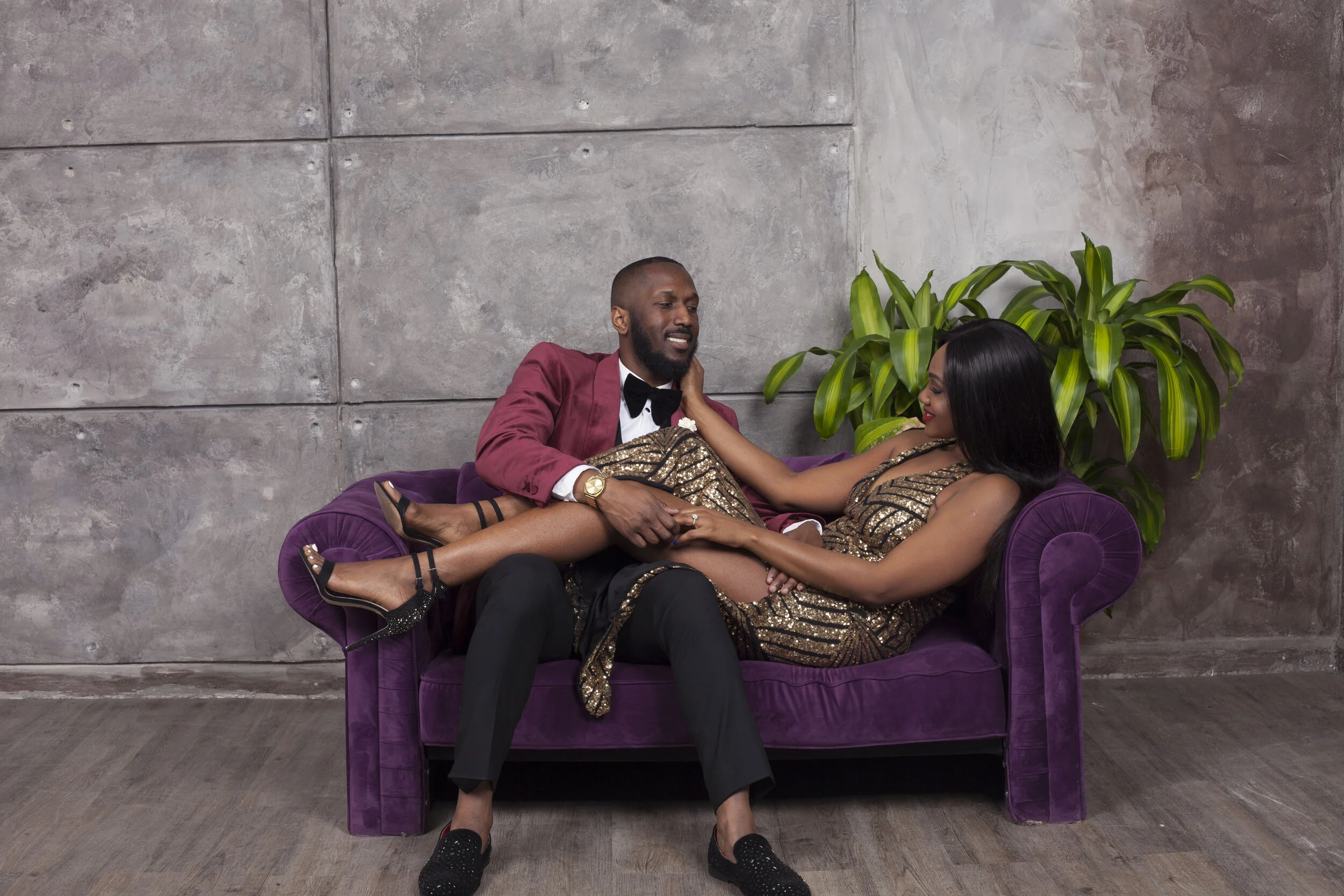 A man and woman sitting on a purple velvet sofa in front of a concrete wall, with a large green plant in the background. The man is wearing a red tuxedo jacket with black bow tie and black pants, and the woman is dressed in a shiny gold and black dre