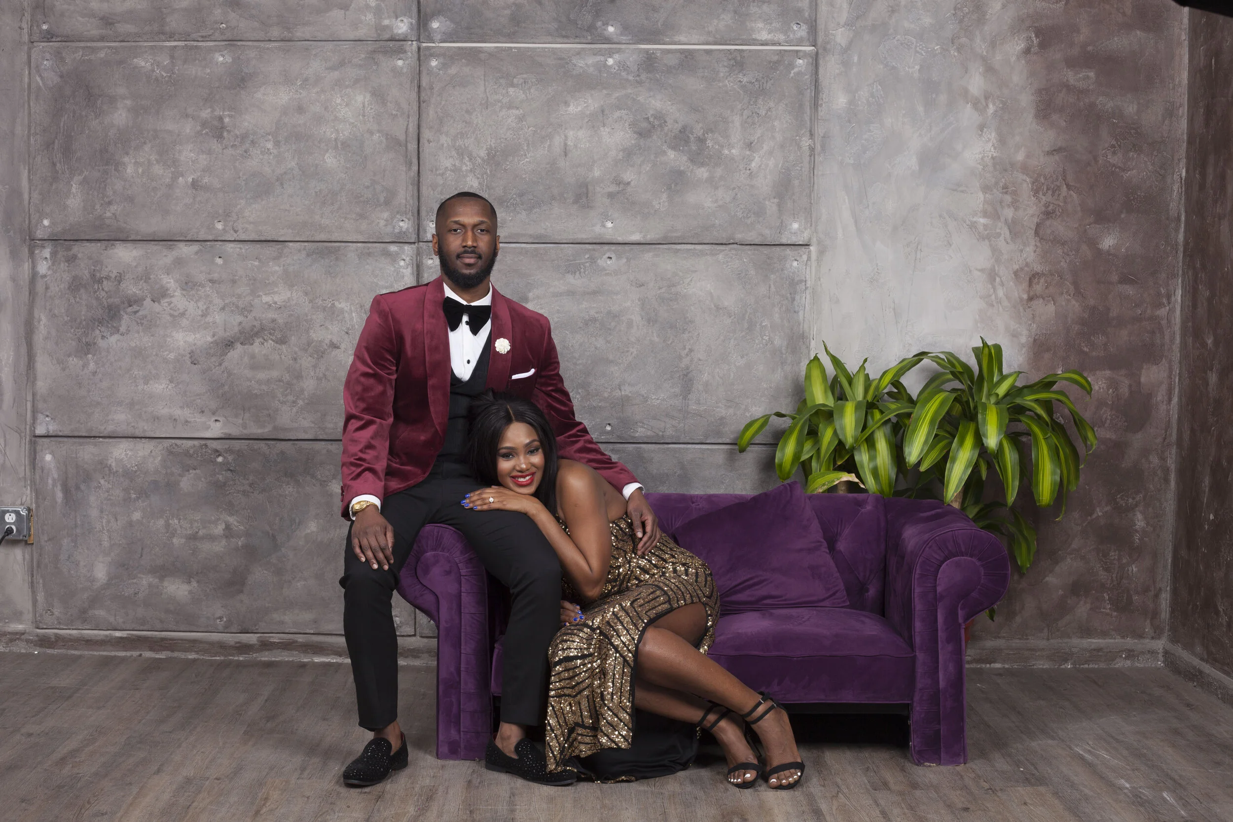 A man in a burgundy tuxedo with a black bow tie standing behind a woman sitting on a purple velvet sofa, with a gray concrete wall in the background and a green houseplant to the right.