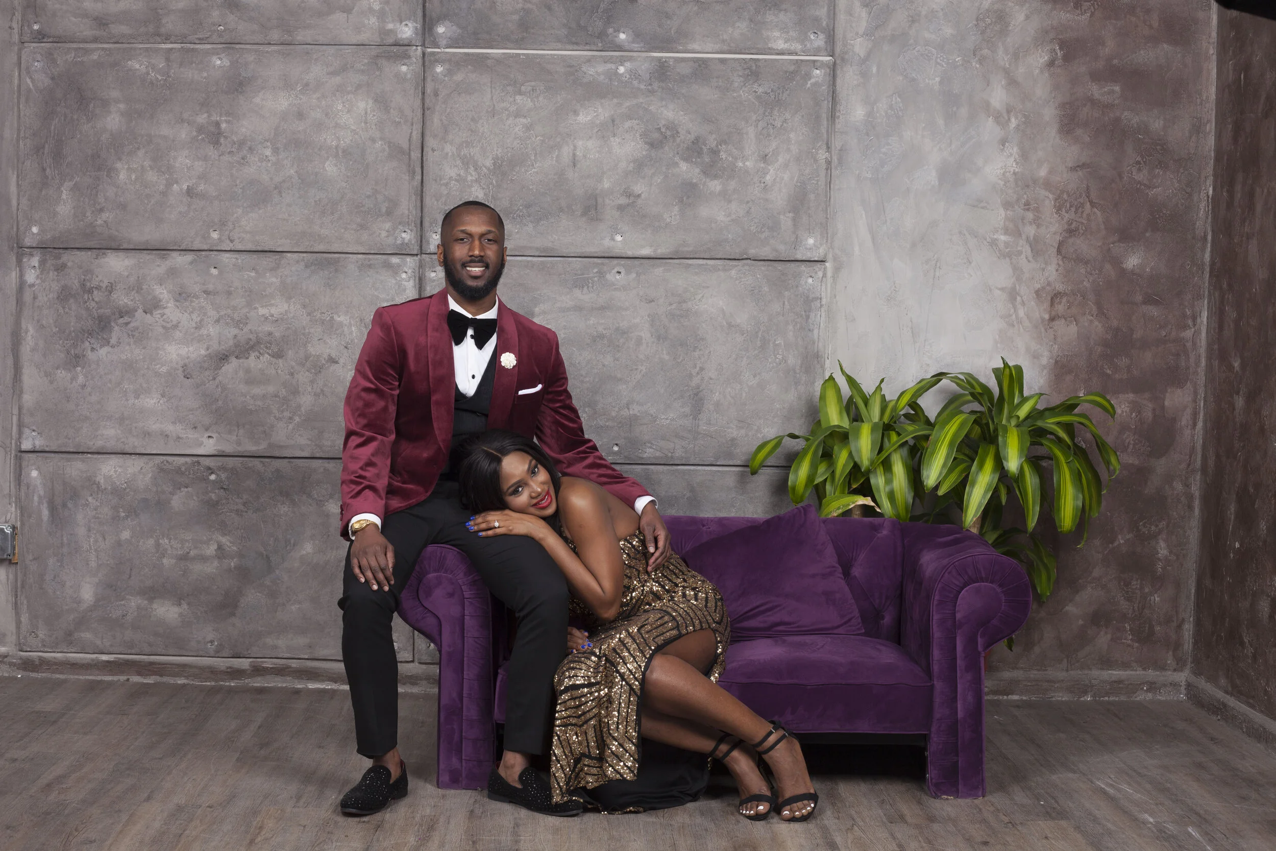 A man and a woman posing together in a studio. The man is standing behind a purple velvet sofa, dressed in a burgundy tuxedo with a white shirt and black bow tie. The woman is lying on the sofa, wearing a gold sequin dress and black high heels. There