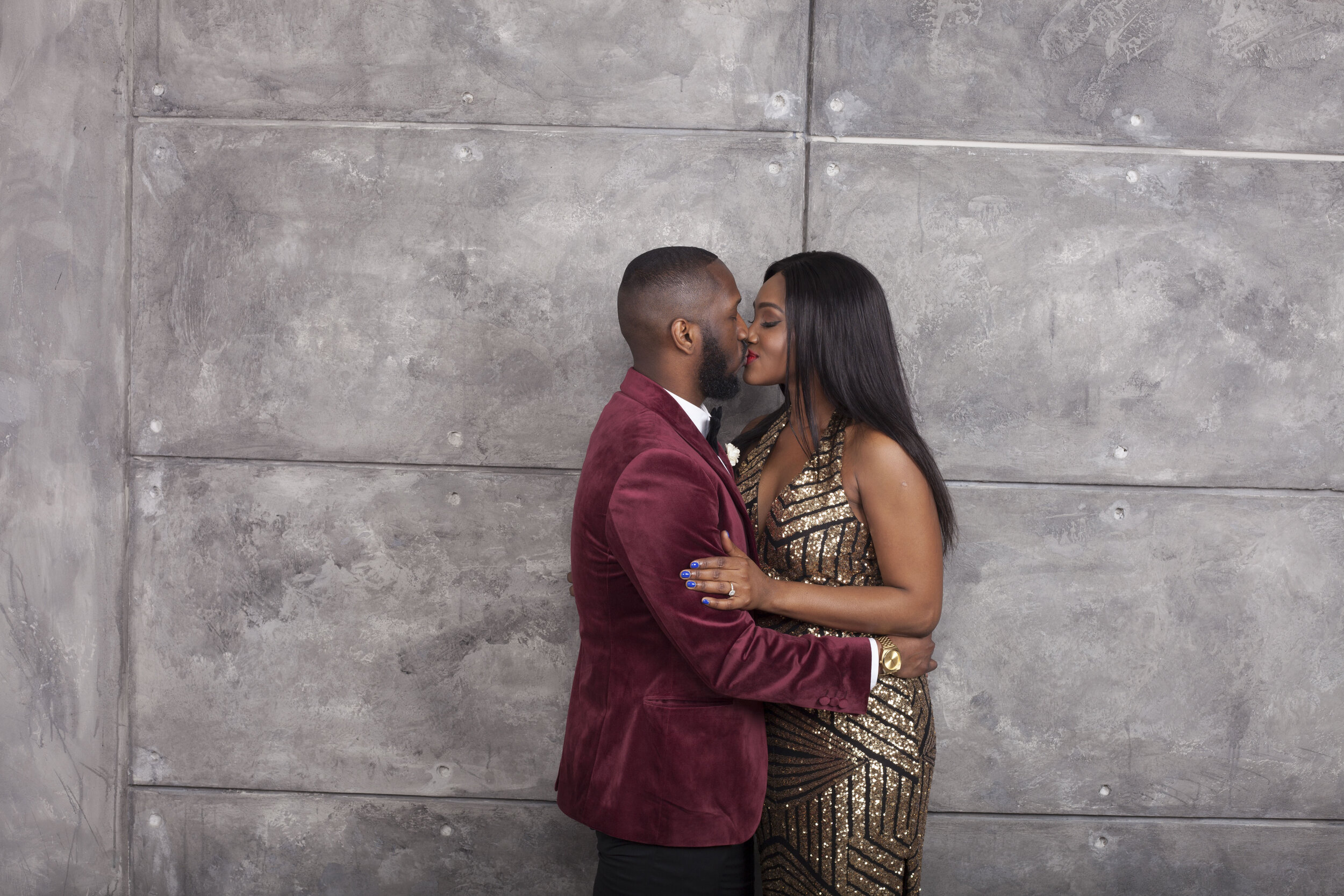 A couple stands close together, about to kiss, against a gray concrete wall. The man wears a red velvet blazer, white shirt, and black bow tie. The woman wears a gold and black sequined dress with long black hair. They are holding each other affectio
