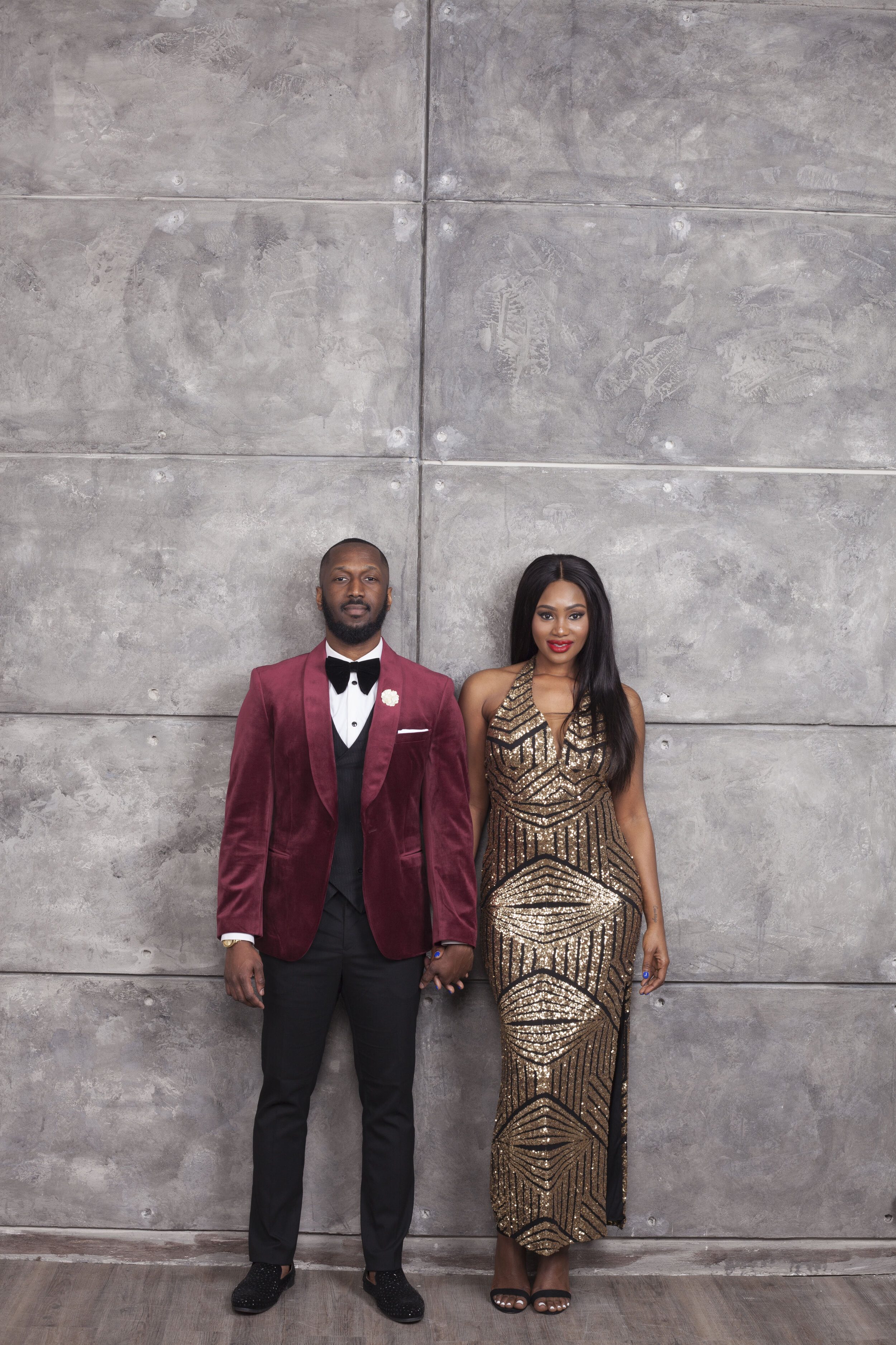 A man and woman standing side by side against a gray industrial-looking wall. The man is wearing a burgundy tuxedo jacket, white shirt, black bow tie, and black pants. The woman is dressed in a glamorous, fitted, gold and black sequined gown with a g