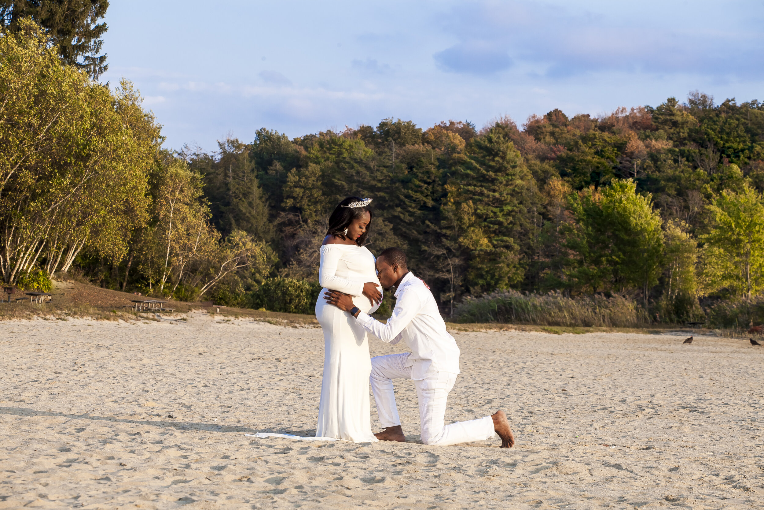 A couple dressed in wedding attire is on a sandy beach, with the man kneeling before the woman who is standing. The man appears to be proposing marriage, and the woman is smiling and looking down at him. There is a lush green forest and a partly clou