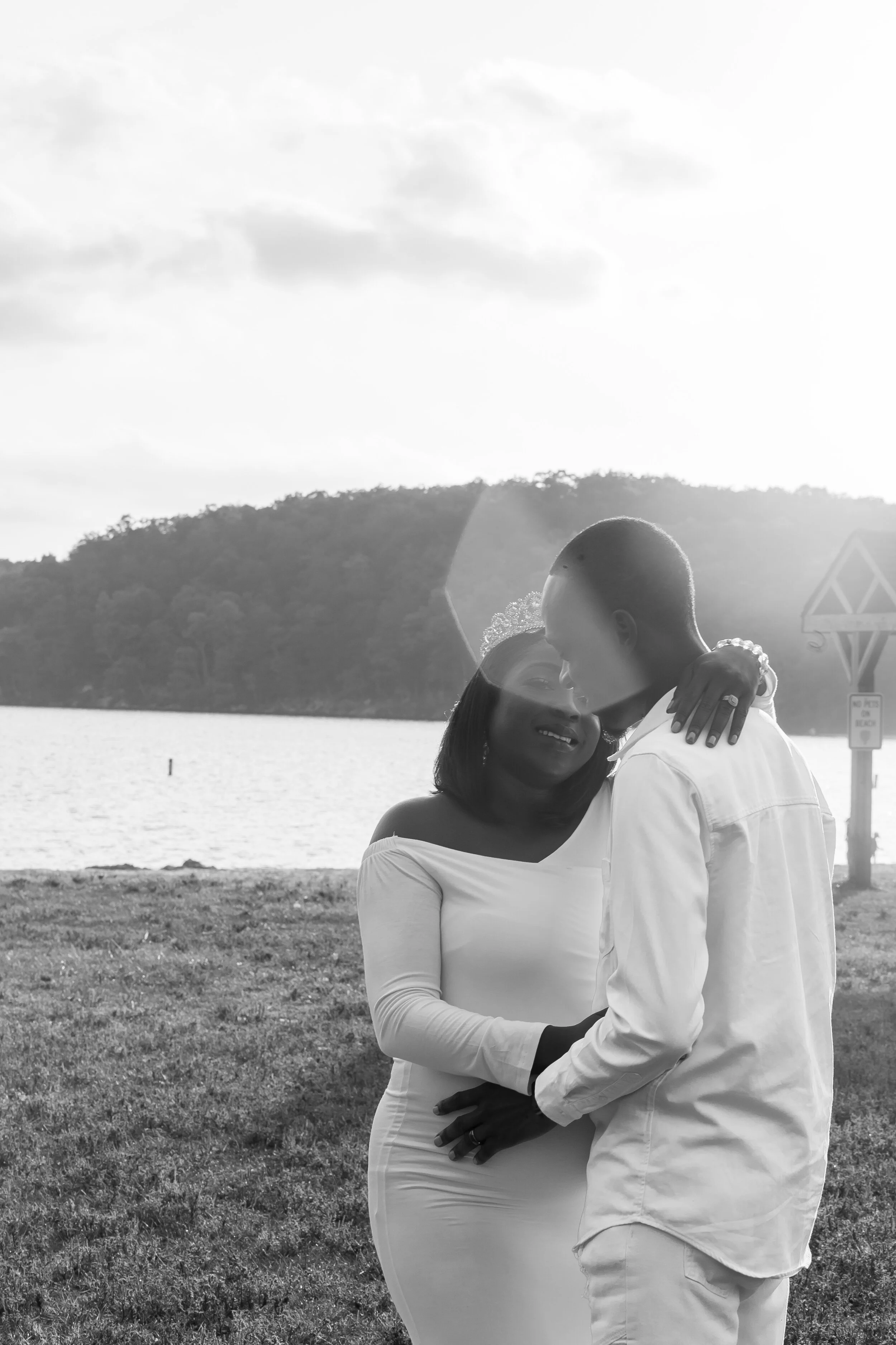 A black-and-white photo of a couple embracing outdoors by a lake, with trees and hills in the background, and a cloudy sky overhead.