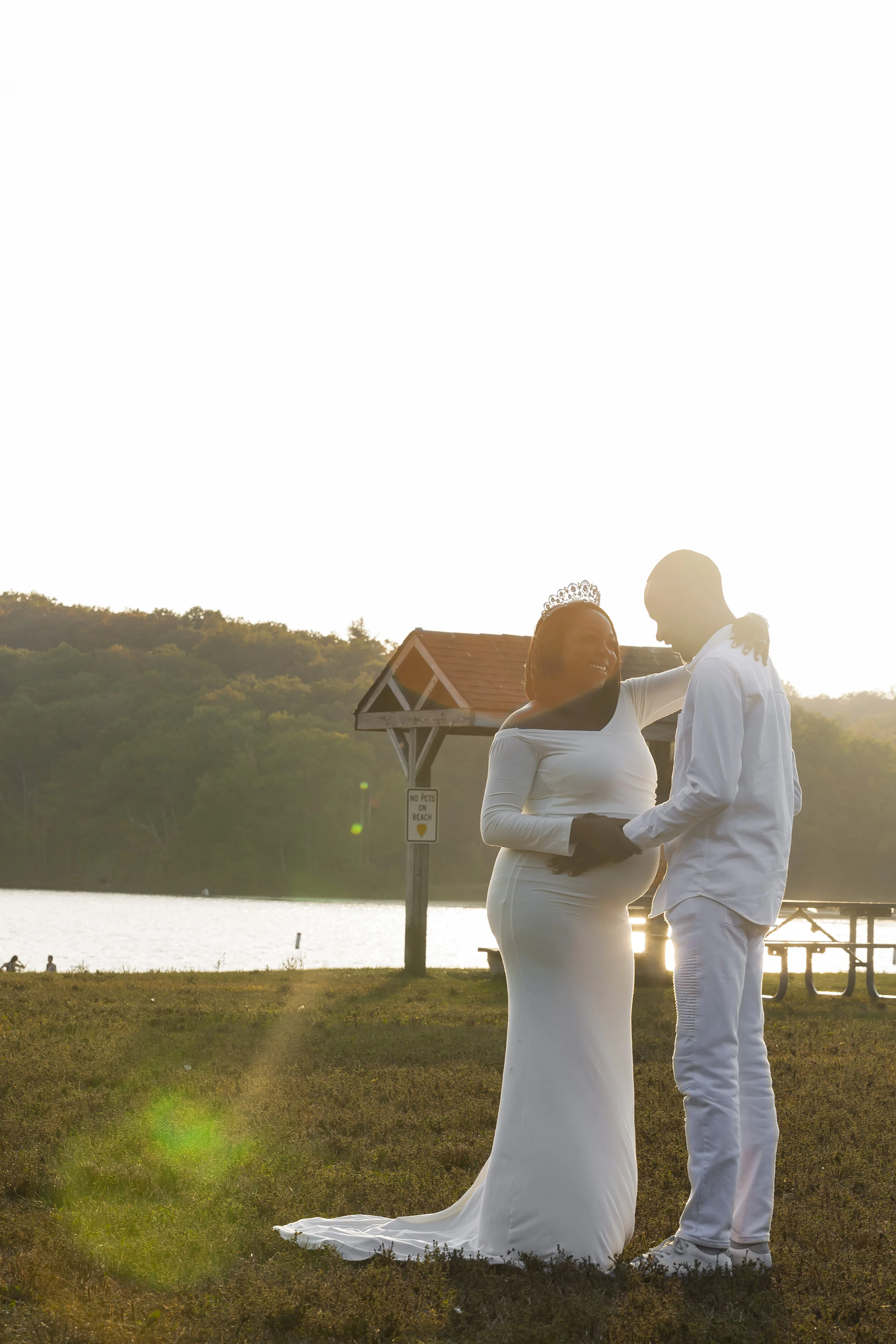 A pregnant woman in a white dress and a tiara smiling at a man dressed in white, standing outdoors near a lake during sunset.