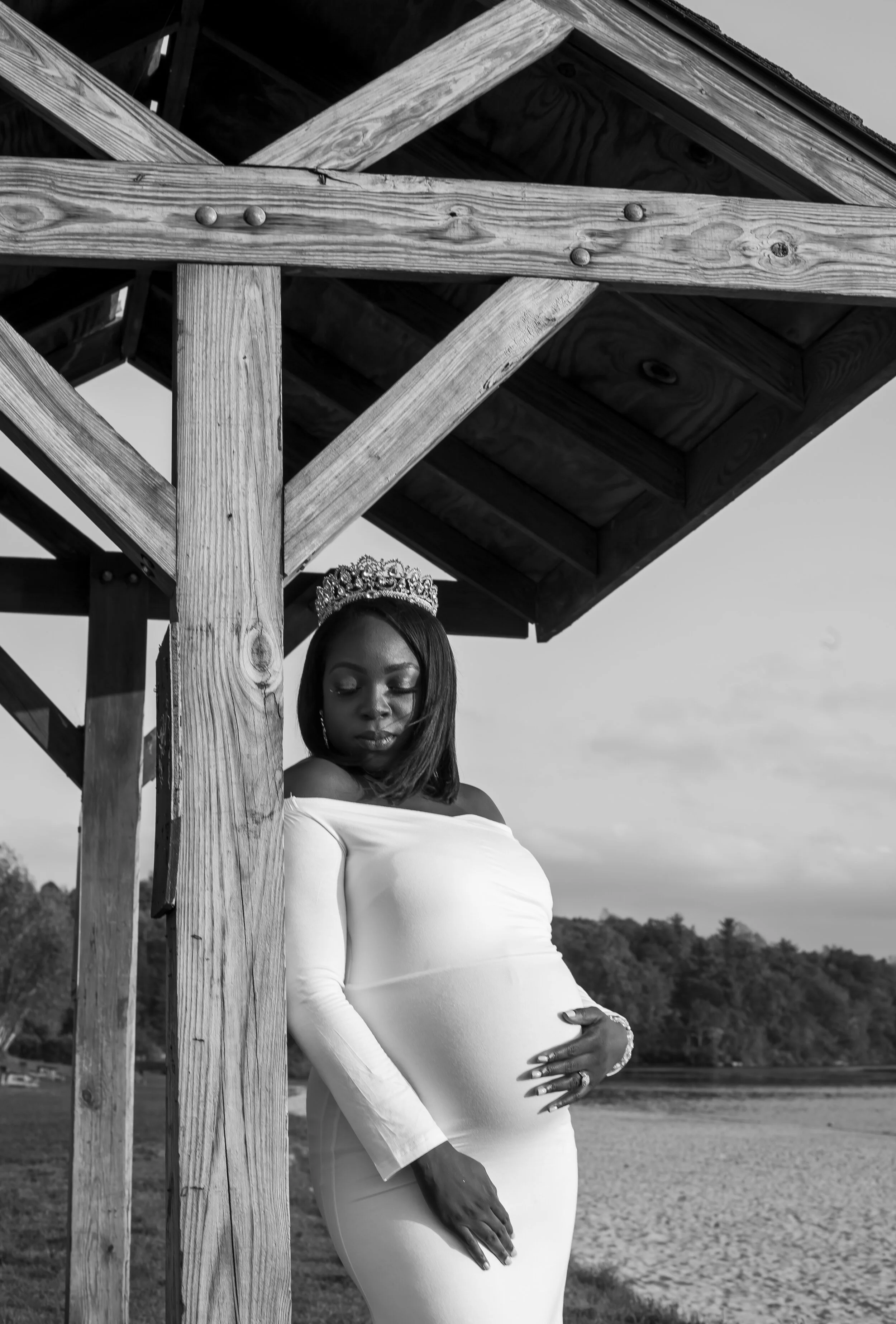 A woman wearing a tiara and a white off-shoulder dress, standing outdoors under a wooden structure, with her eyes closed and one hand on her pregnant belly.