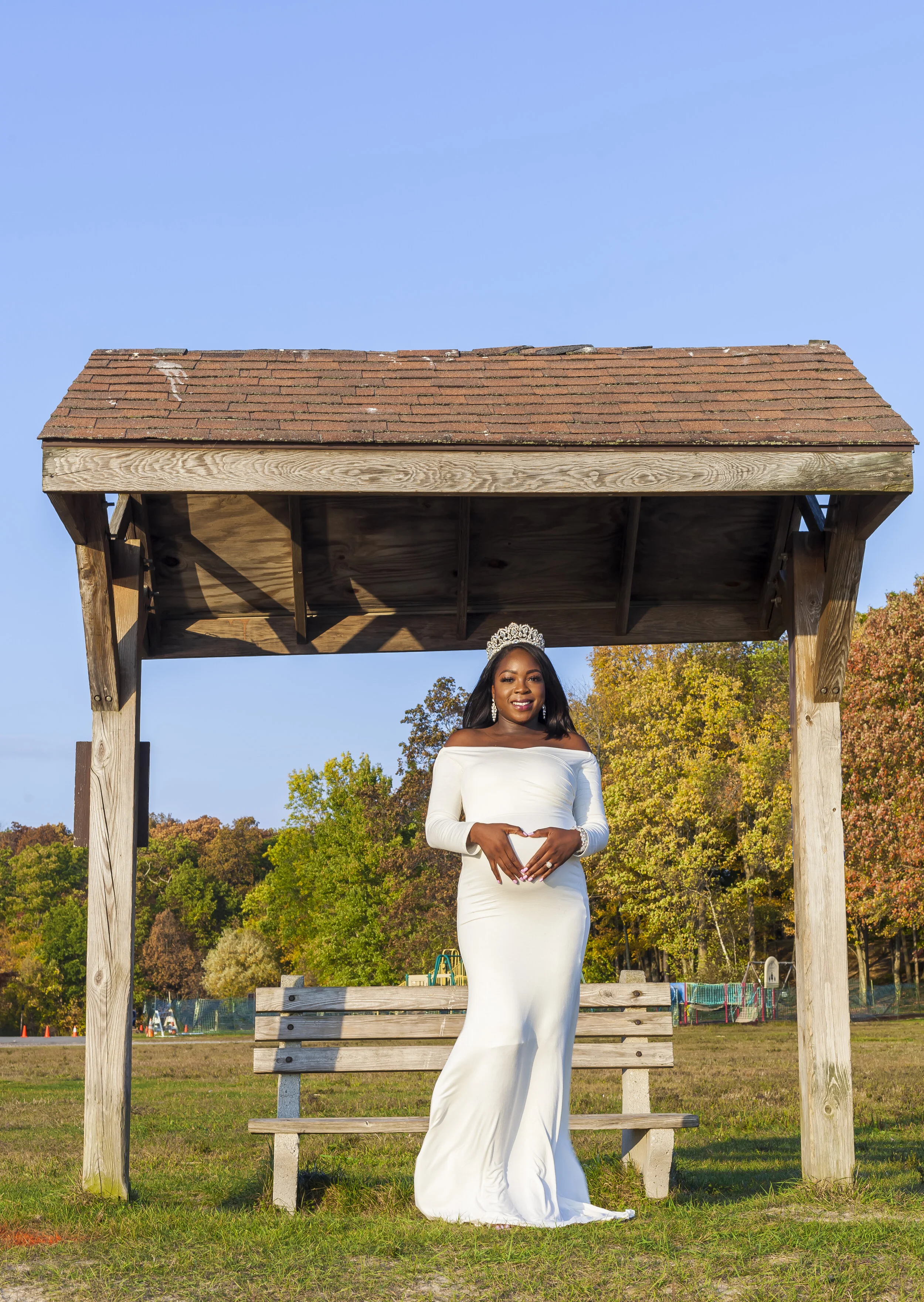 A woman in a white off-shoulder dress and tiara standing under a wooden shelter in a park with green trees and a clear blue sky.