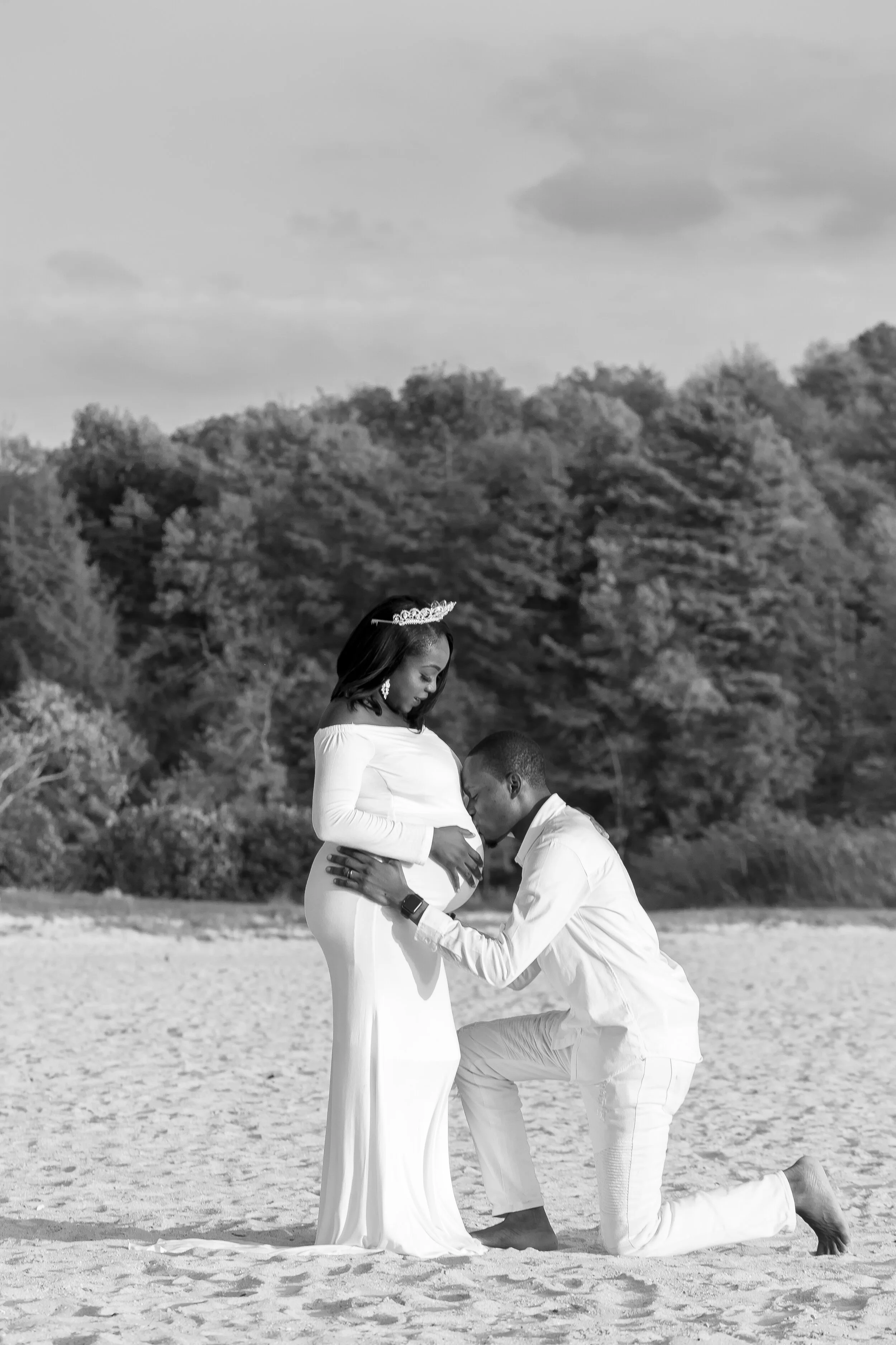 A pregnant woman in a long white dress and tiara standing on a beach as a man kneels before her, kissing her belly, with outdoor trees and cloudy sky in the background.