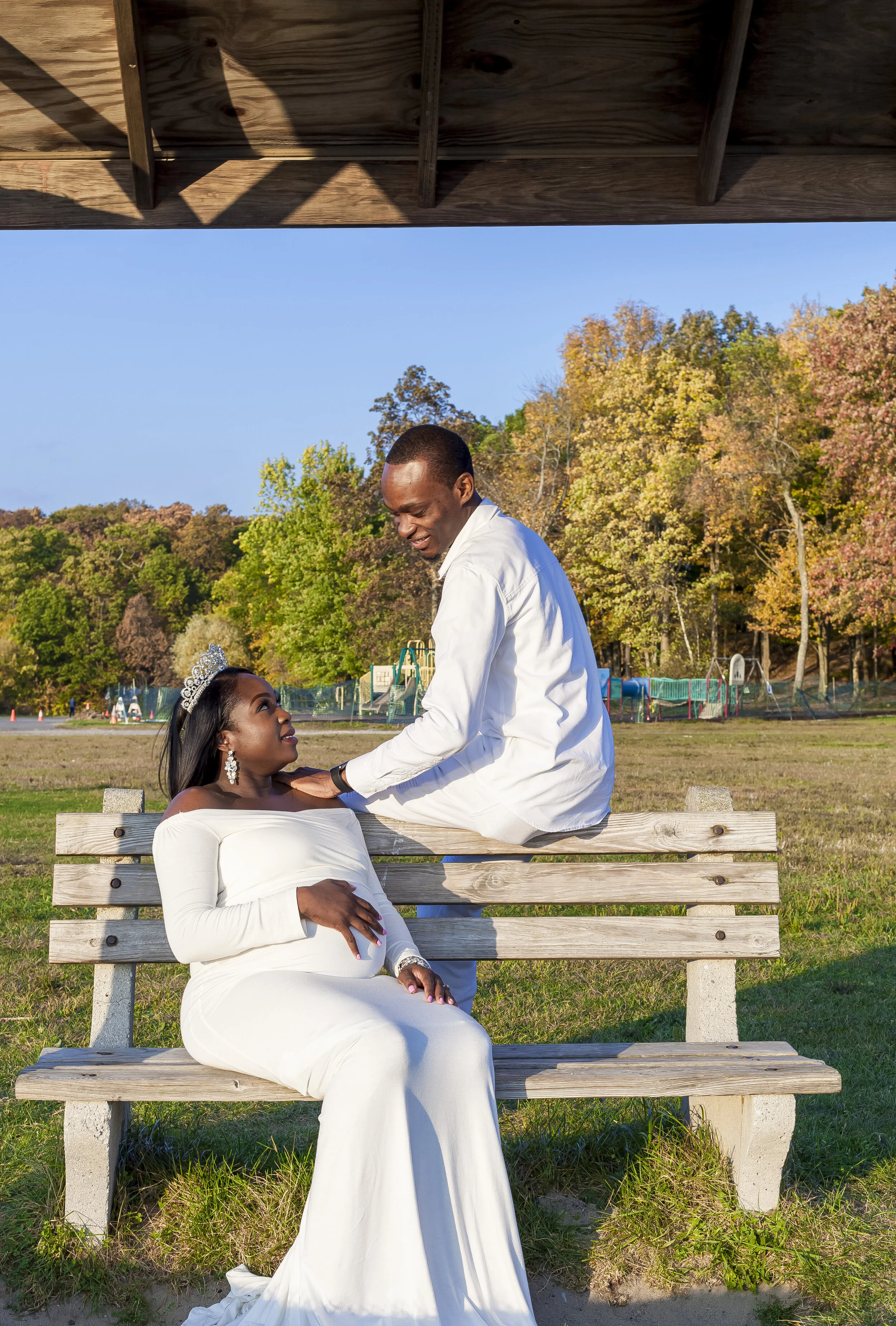 A woman in a white dress and tiara sits on a wooden bench, looking up at a man who is kneeling in front of her with his hand on her shoulder. They are in an outdoor park with trees and playground equipment in the background, and the sky is blue with 