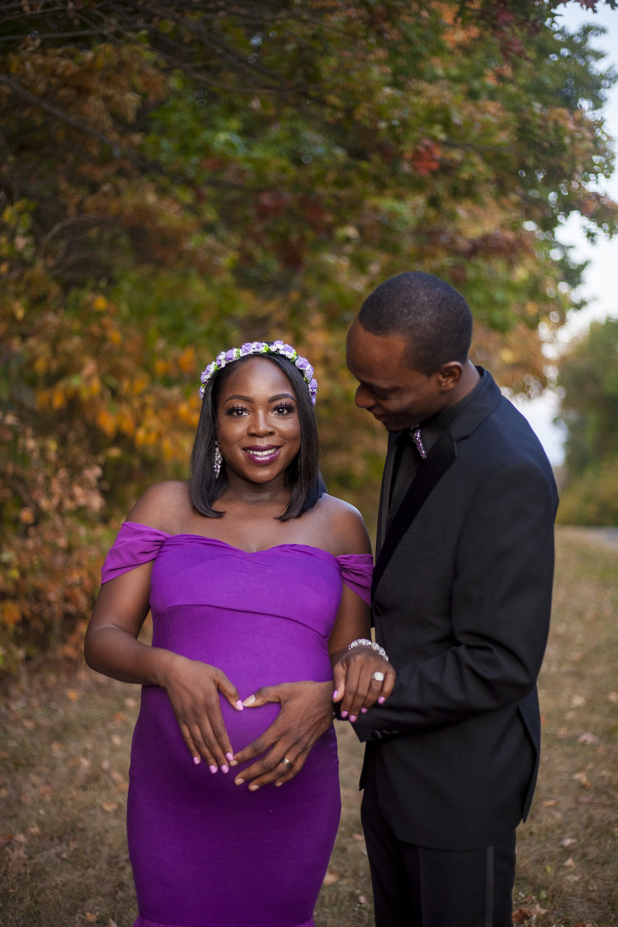 A woman in a purple off-shoulder dress with a floral headband and a man in a black suit with a bow tie standing together outdoors with autumn foliage in the background, holding hands