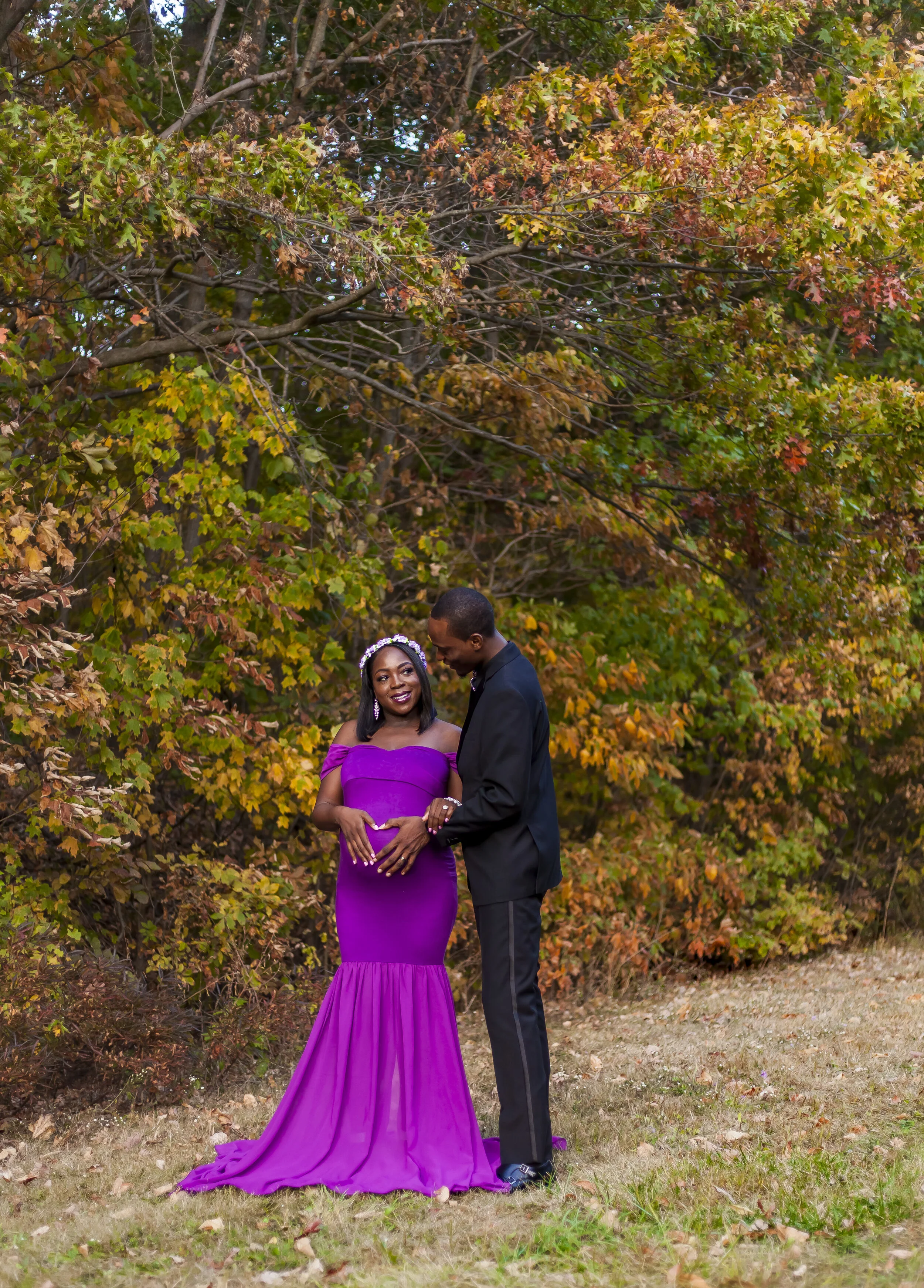 A pregnant woman in a strapless purple gown with ruffled hem standing next to a man in a black suit, outdoors among autumn trees with orange and green leaves, both smiling.