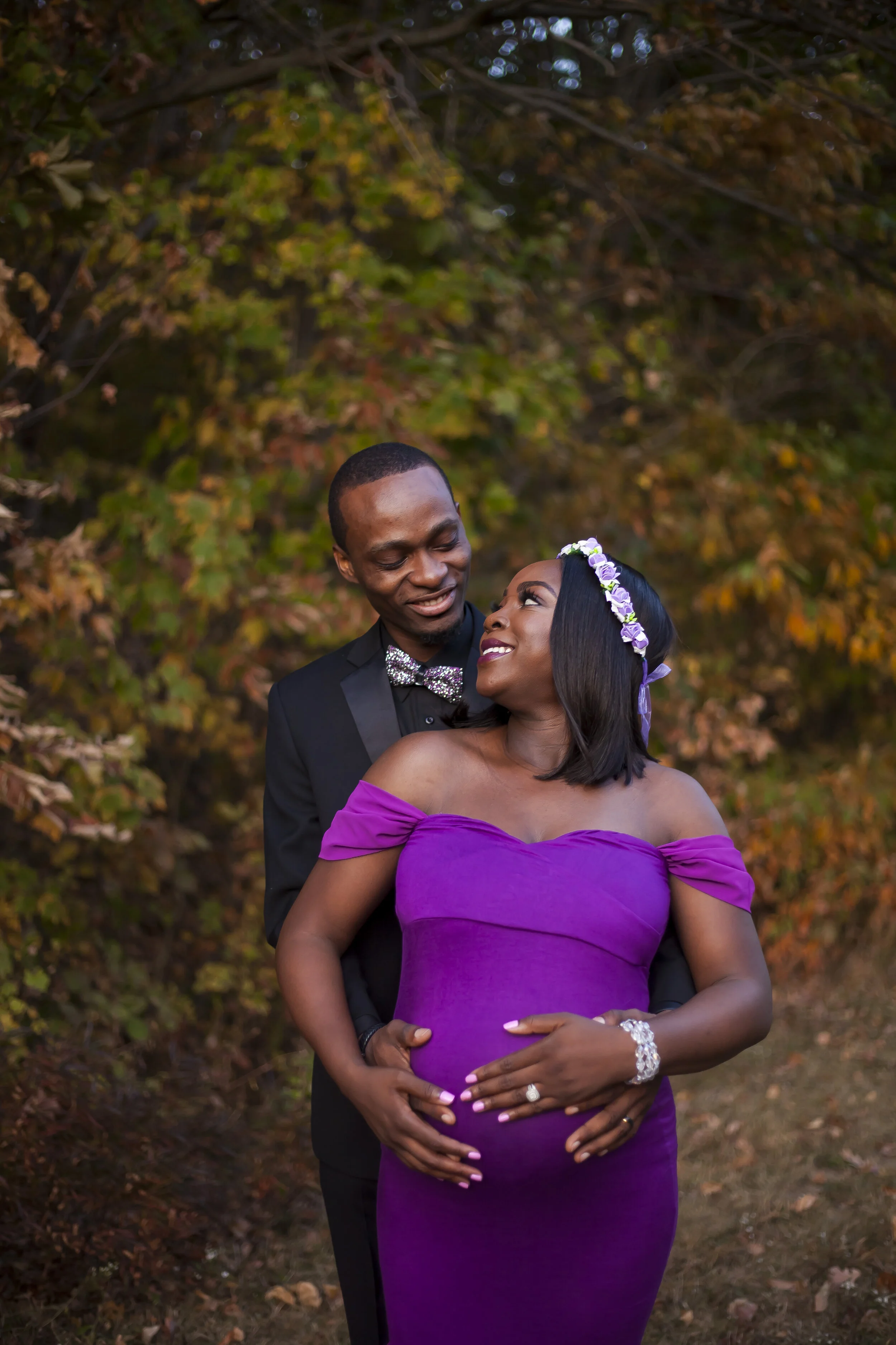 A happy couple standing outdoors, the man in a black tuxedo with a bow tie, and the woman in an off-the-shoulder purple dress, wearing a floral headband, during autumn.