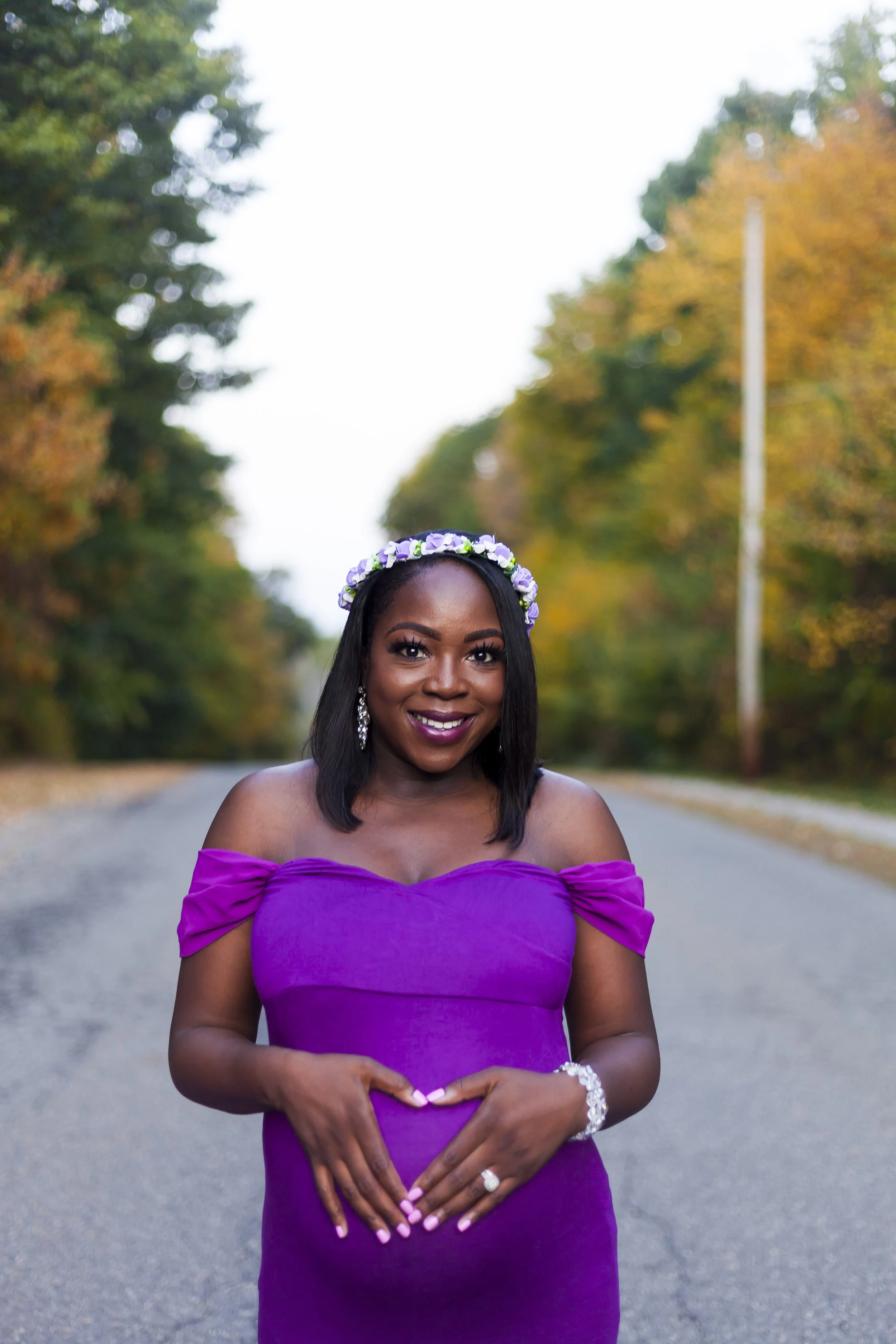 A smiling woman in a purple off-the-shoulder dress with a flower crown on her head, standing on a tree-lined road during autumn.