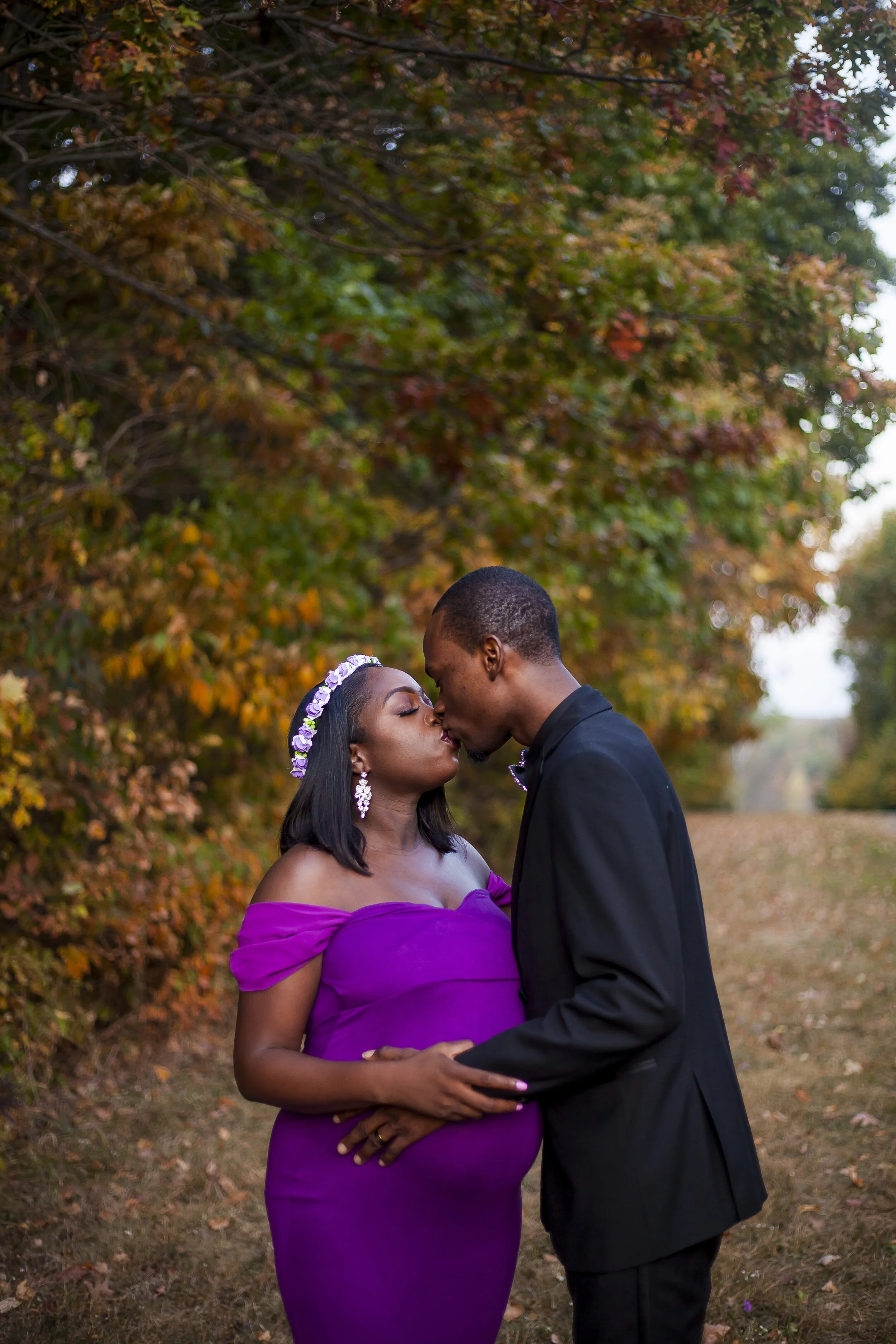A couple is standing outdoors in an autumn setting, with colorful fall leaves on trees behind them. They are close, touching foreheads, with eyes closed and gentle expressions. The woman is wearing a purple off-shoulder dress and a floral headband, a