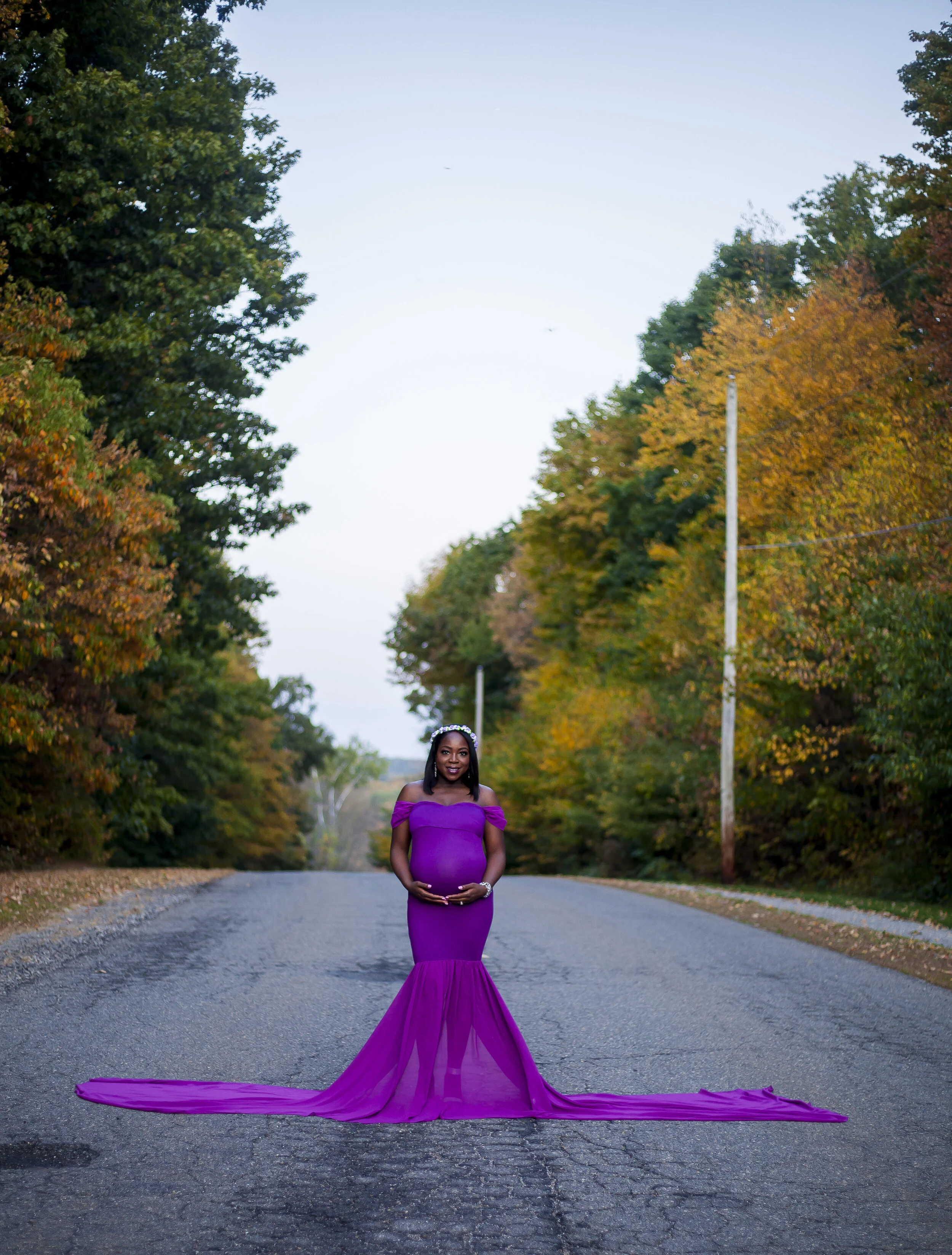 A pregnant woman in a purple gown standing on an empty road surrounded by trees with fall foliage, holding her belly.
