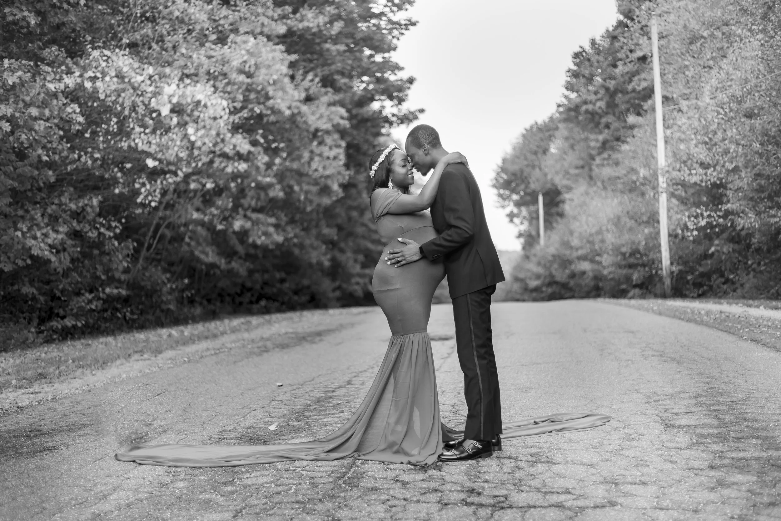 A couple standing on a road surrounded by trees, embracing each other as the woman wears a long gown and the man is dressed in a suit. They are holding each other's faces close with foreheads touching.
