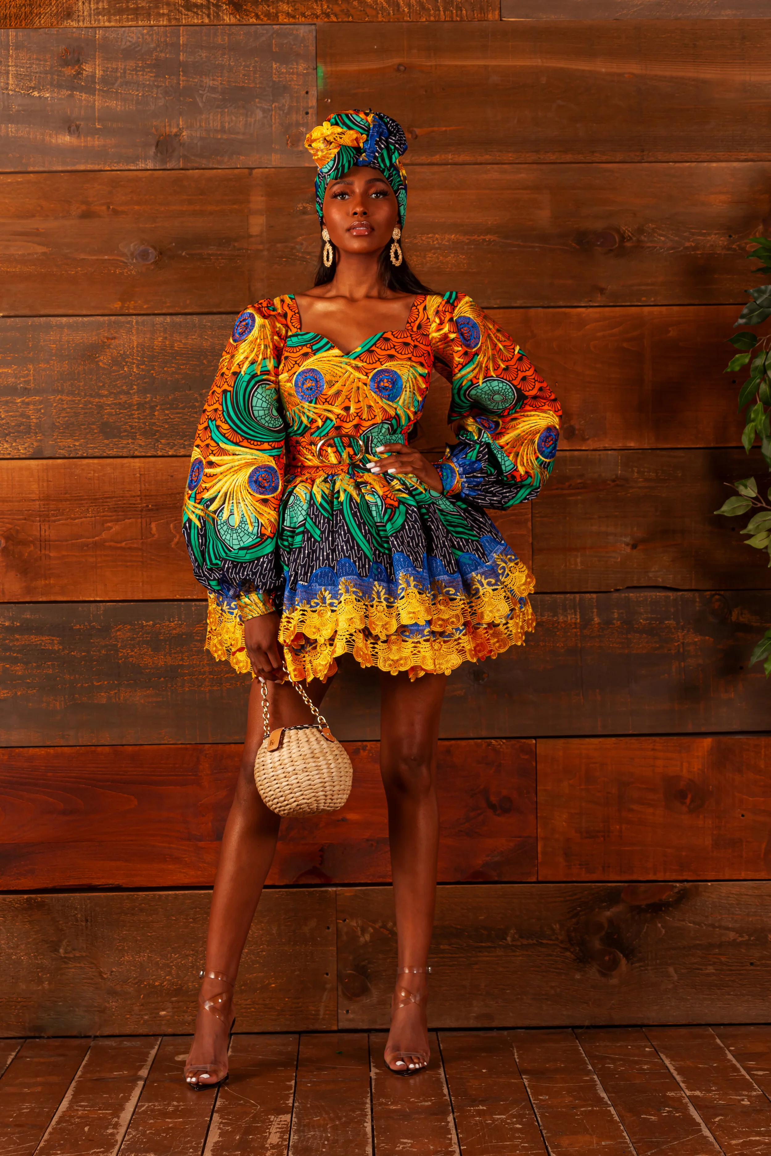 A woman standing with her hand on her hip against a wooden wall. She is wearing vibrant, colorful African-print clothing and a matching headwrap, accessorized with earrings and holding a small woven handbag. She is wearing clear high heels.
