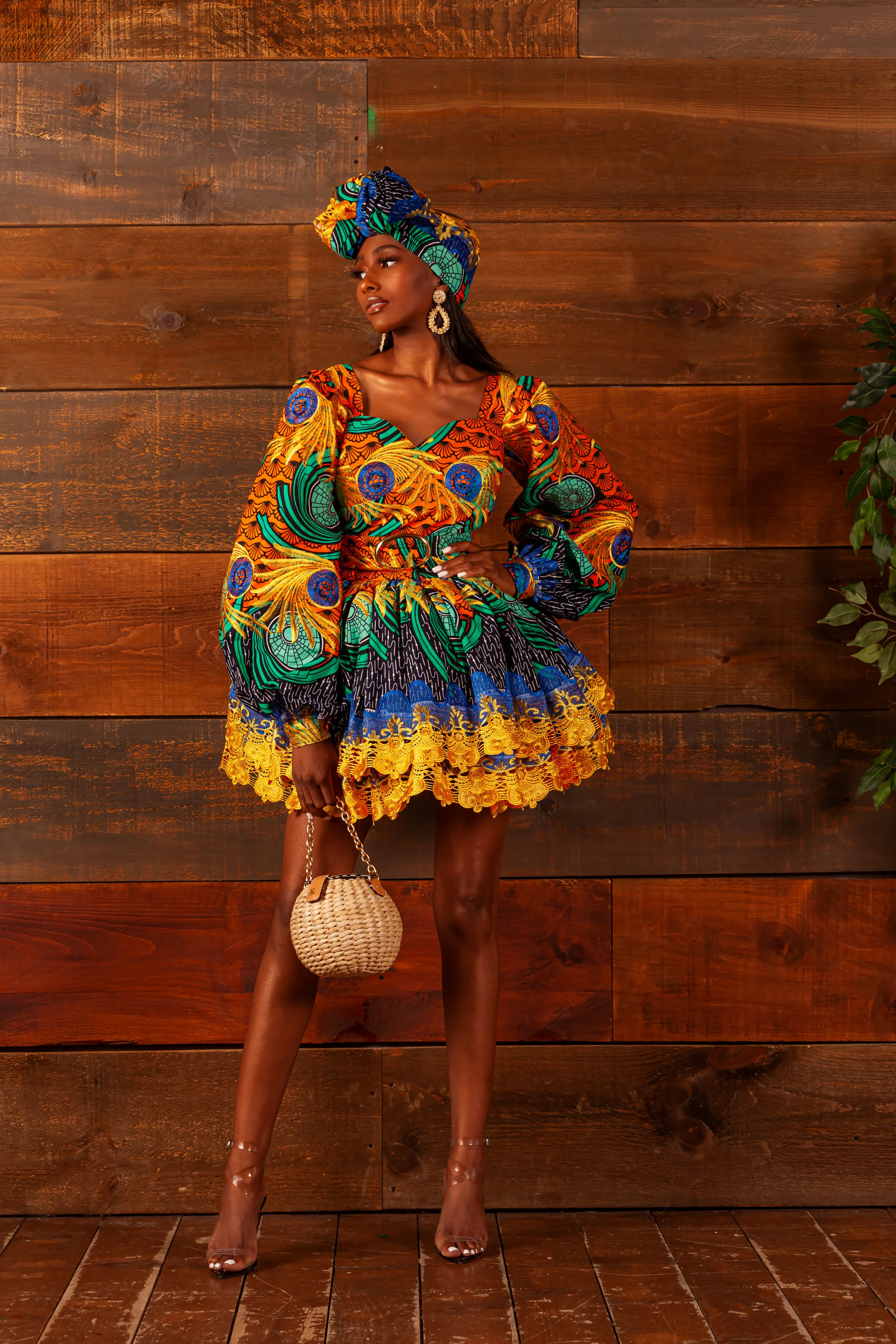 A woman standing against a wooden wall, wearing a colorful African print dress with puffed sleeves and a matching headwrap. She is holding a small woven handbag and wearing high heels and large earrings.