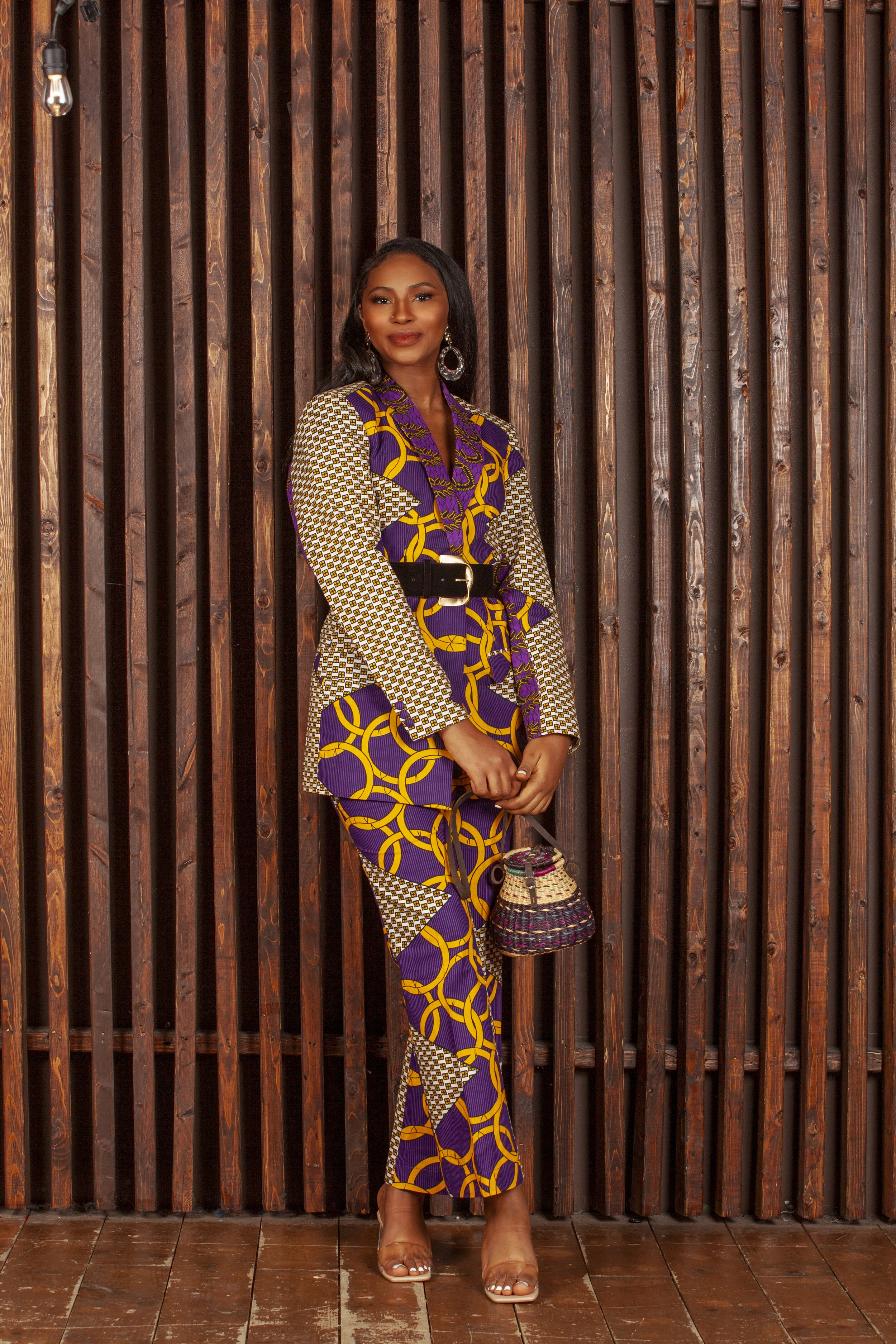 A woman in a colorful, patterned outfit stands against a wooden wall, holding a woven handbag, and wearing large hoop earrings and clear heels.