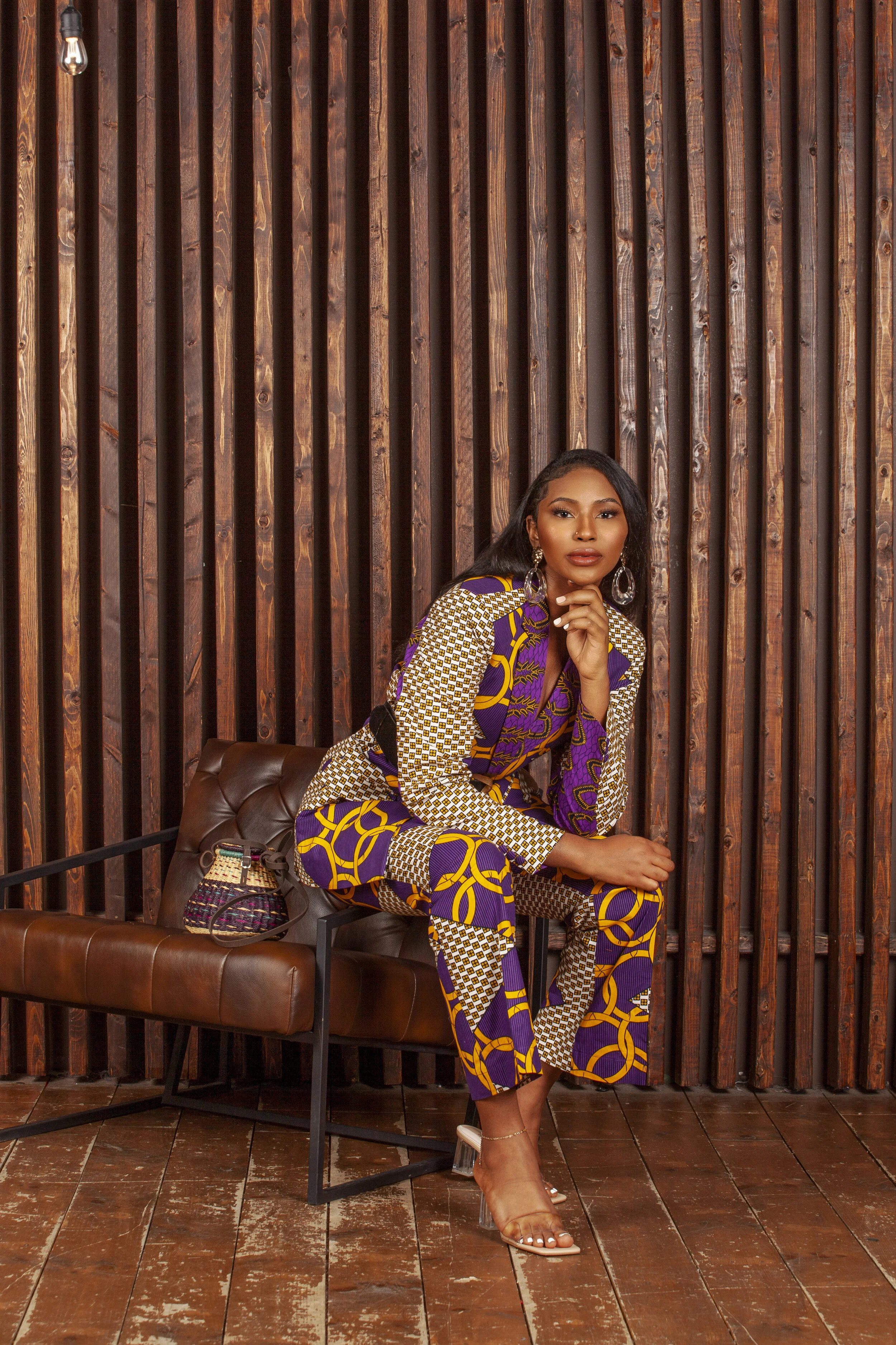 A woman with dark hair and medium skin tone seated on a brown leather bench in front of a wooden wall, wearing colorful patterned clothing and clear high heels, with a small purse beside her.