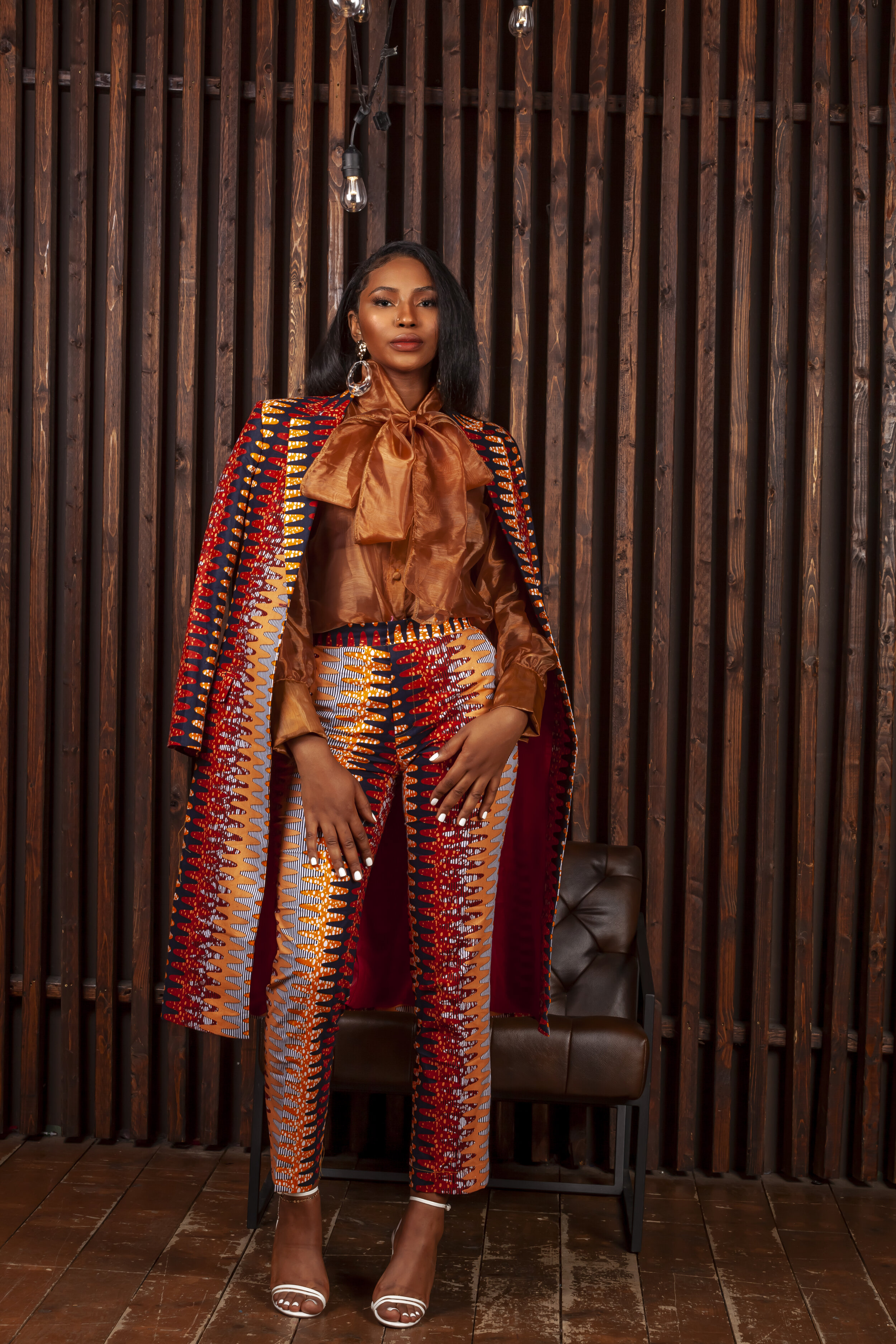 A woman standing in front of a dark wooden slat wall, wearing a colorful patterned pantsuit with matching cape, a shiny bronze blouse with a bow tie, and white high-heeled sandals.