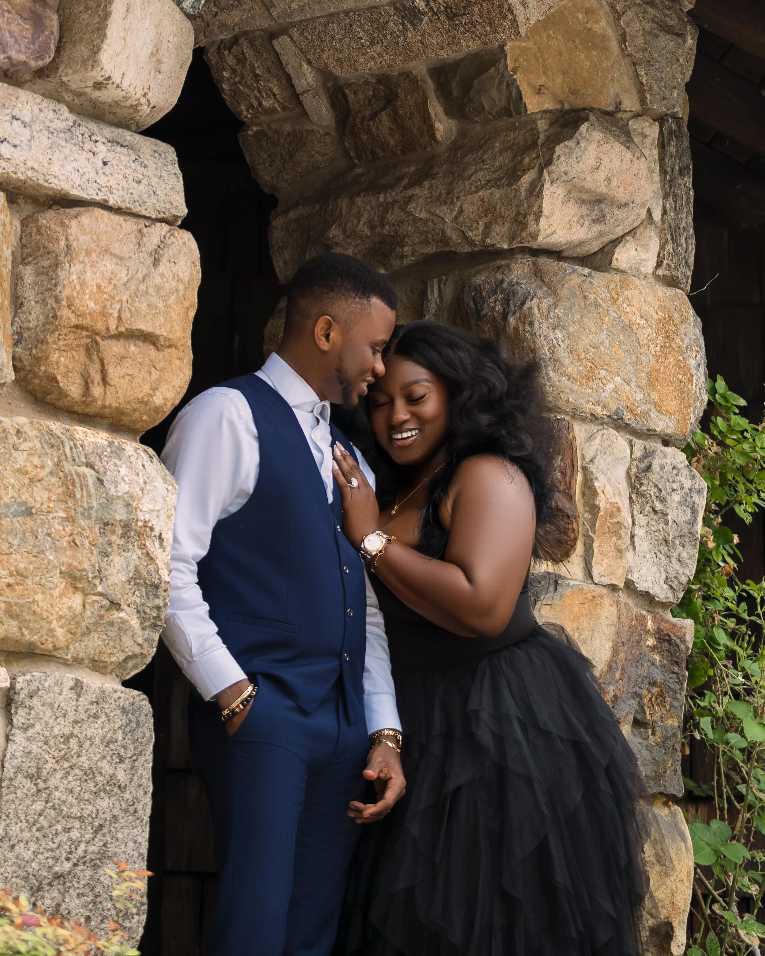 A couple is standing close together, smiling and with foreheads touching, in front of a stone wall. The man wears a white shirt and blue vest, and the woman wears a black strapless dress and jewelry.