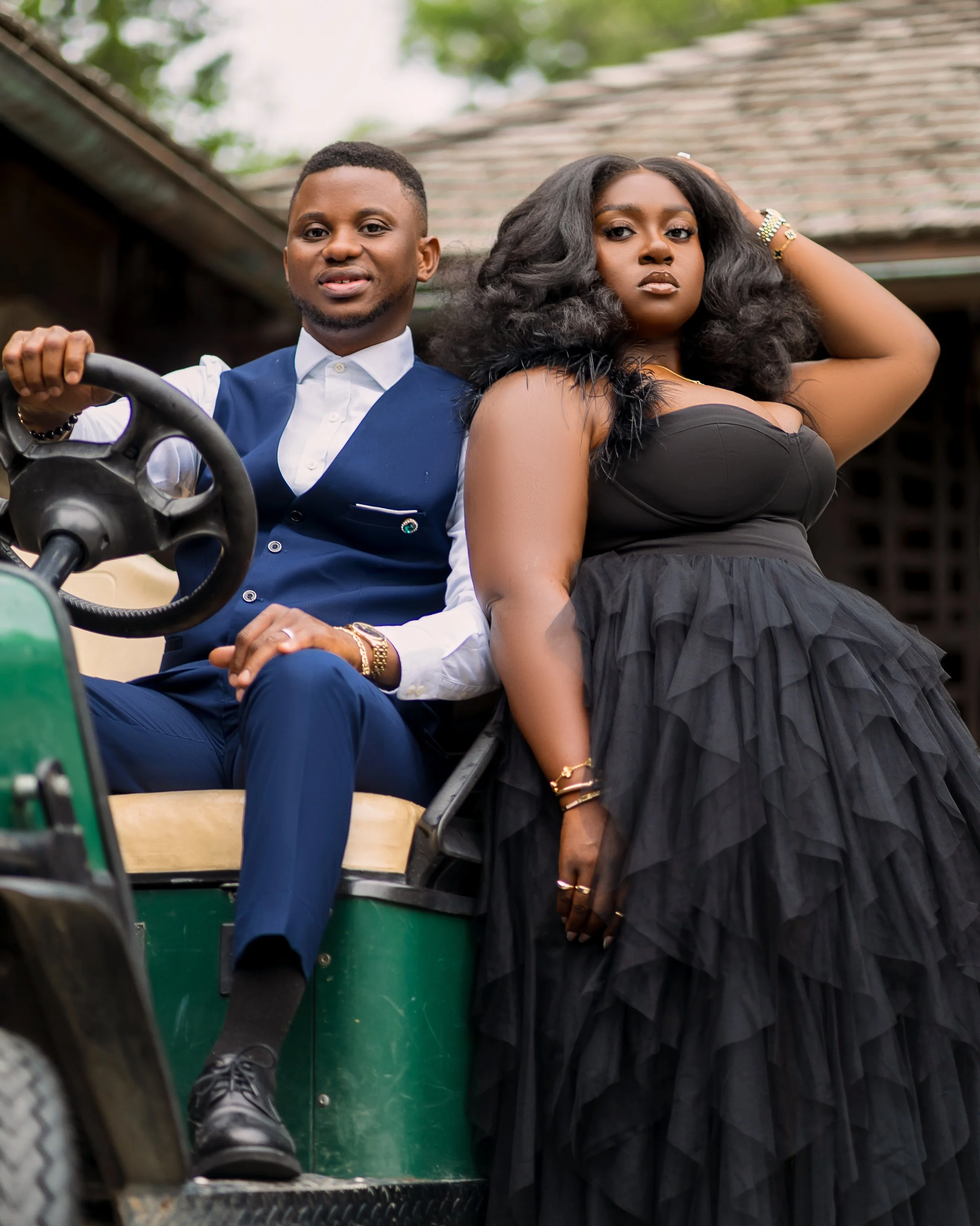 A man and woman sit together on a green open-air vehicle, looking at the camera. The man is holding a steering wheel and wears a white shirt with a blue vest, while the woman has voluminous black hair, gold bracelets, and a strapless black dress.